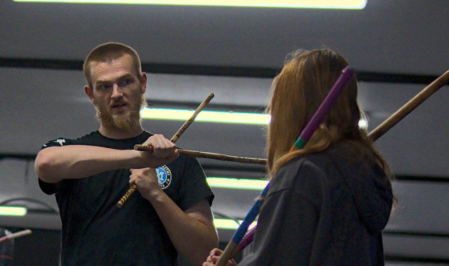 A martial arts instructor demonstrates stick fighting technique with a student indoors.