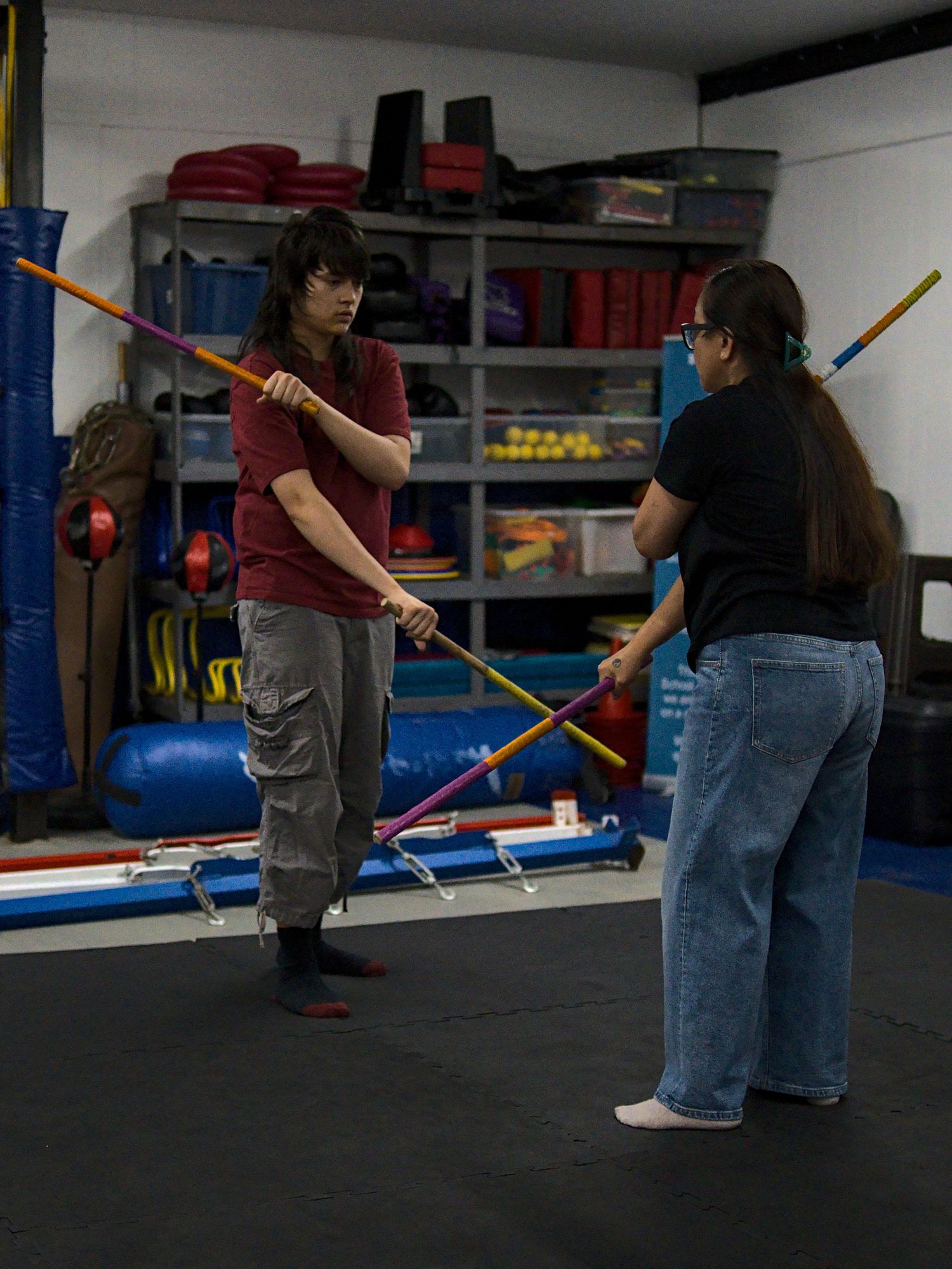 Two students practice Filipino martial arts stick fighting techniques in a well-equipped indoor training gym.