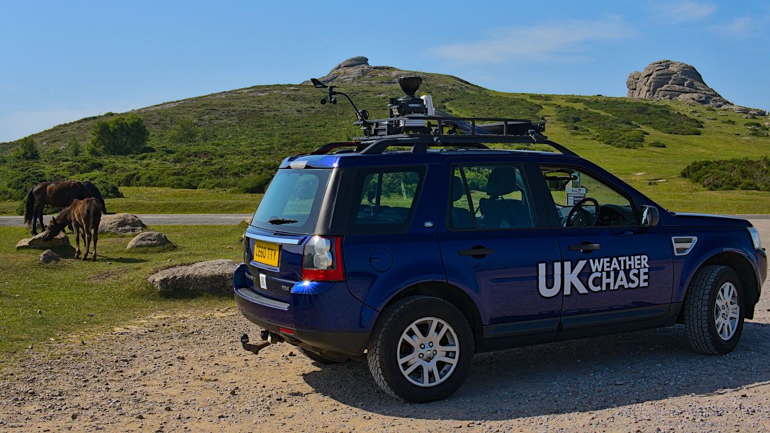 UK Weather Chase blue SUV parked on rocky moorland with horses grazing near dramatic rocky tor landscape.