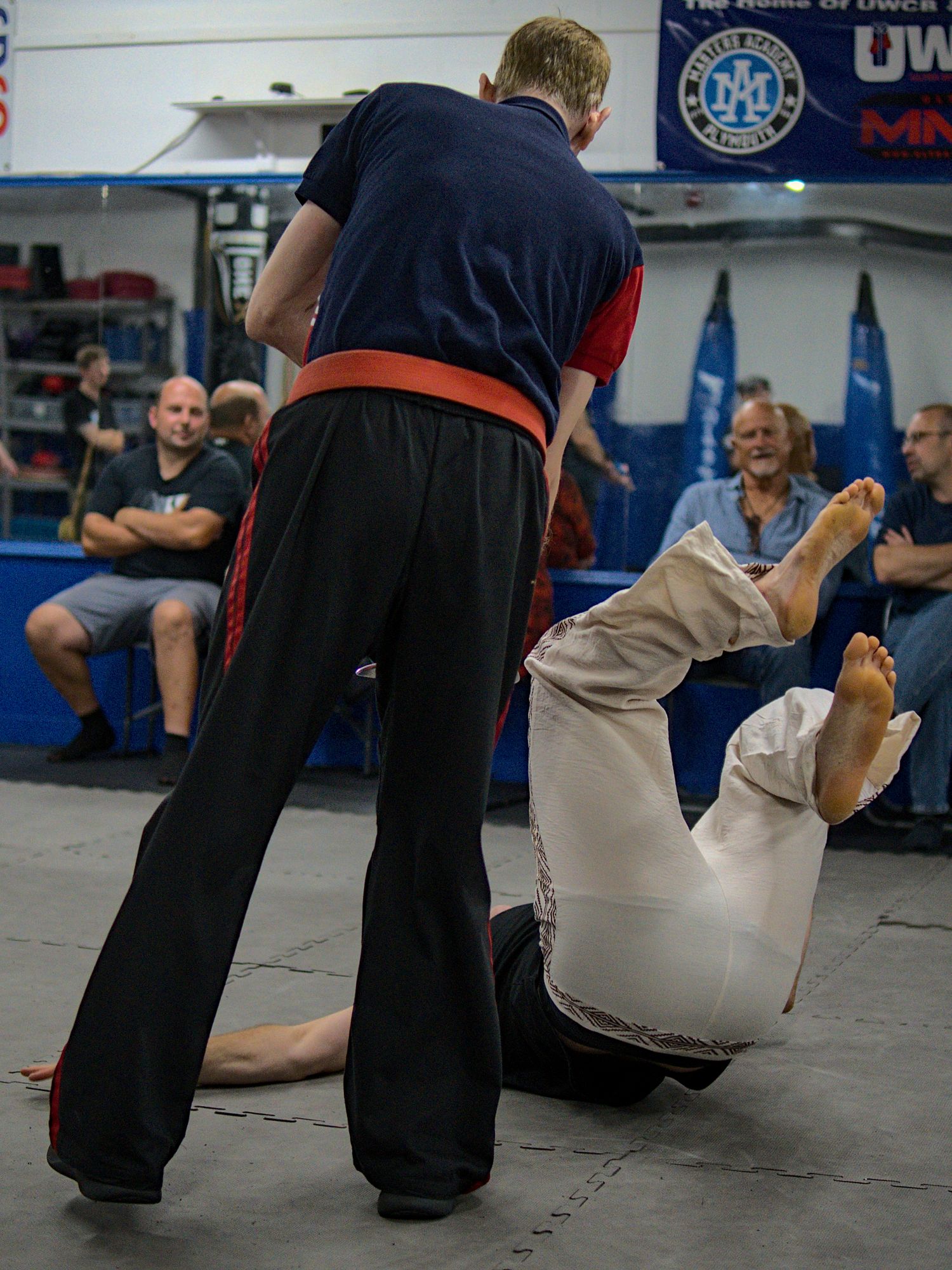 Martial arts instructor stands over fallen student demonstrating takedown technique at indoor training facility.
