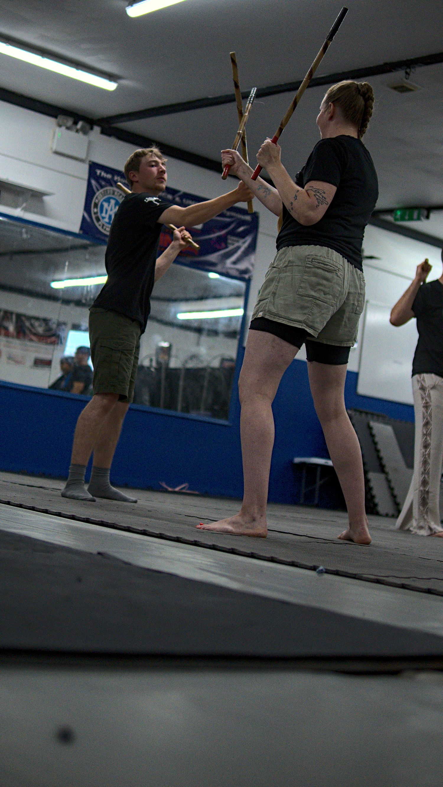 Two students practice stick fighting techniques with rattan sticks in a martial arts gym.