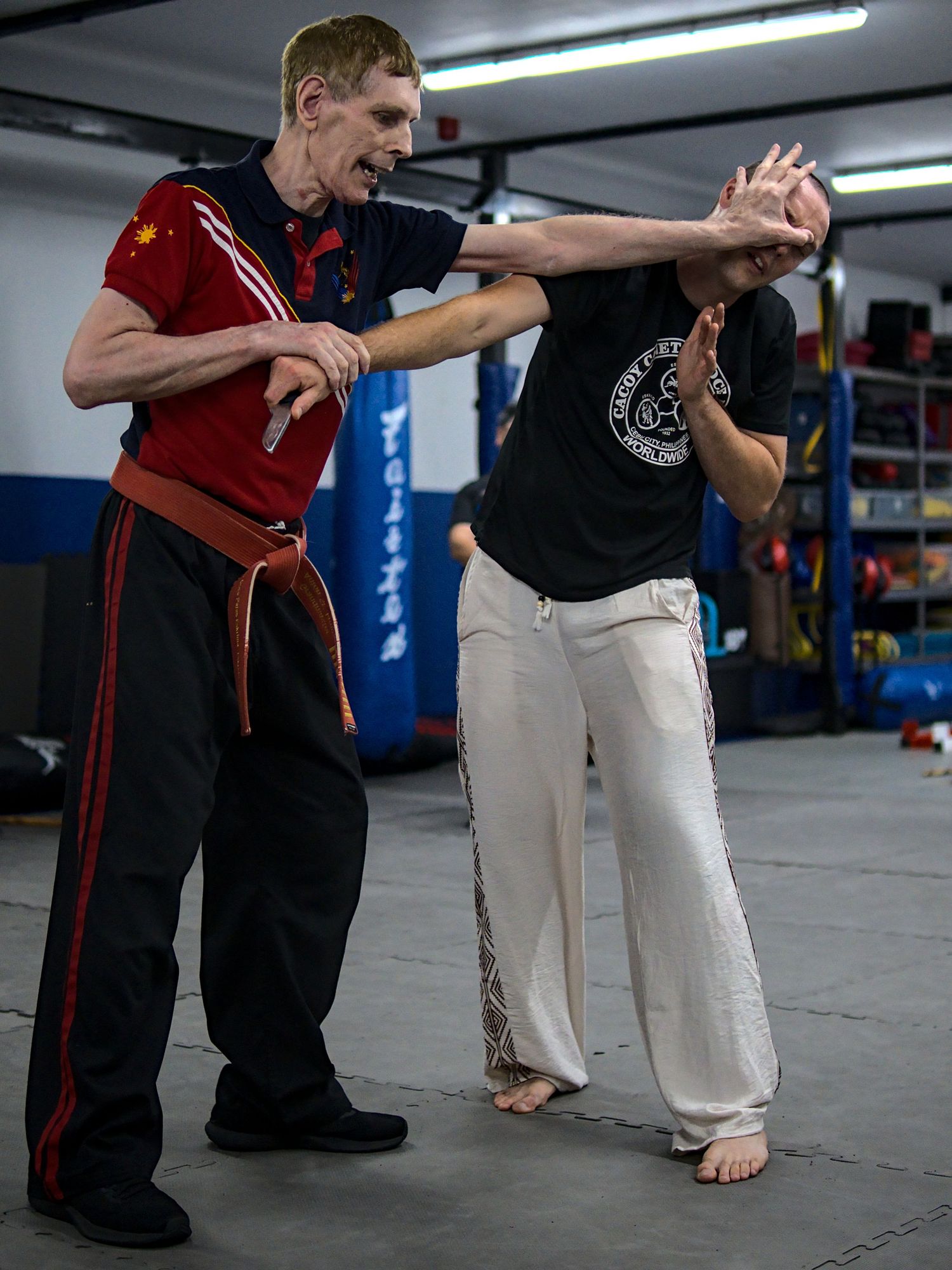 Martial arts instructor in red uniform demonstrates knife defense technique with student in black shirt at indoor dojo.