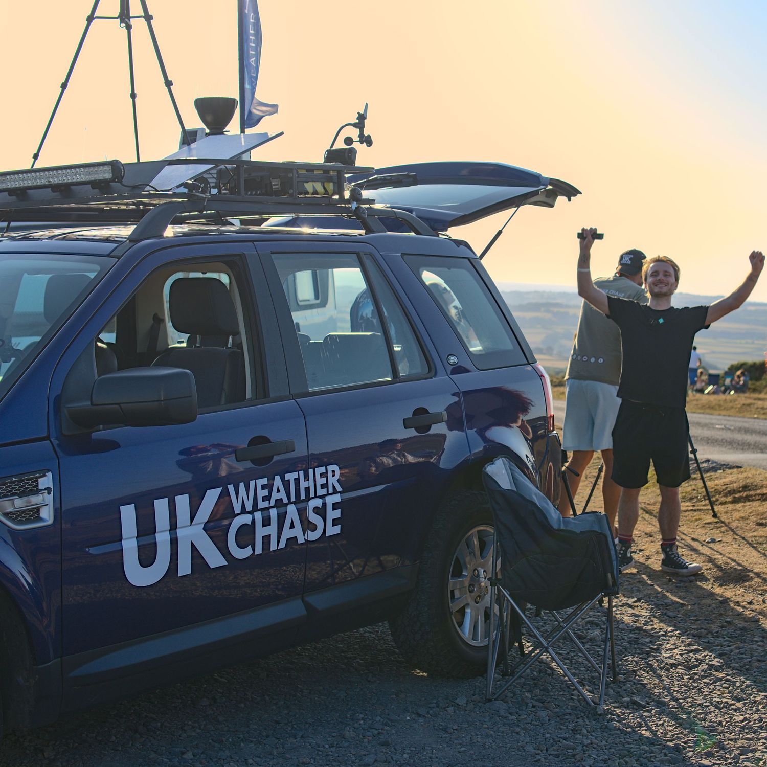 Man celebrating with arms raised next to UK Weather Chase vehicle during golden sunset.
