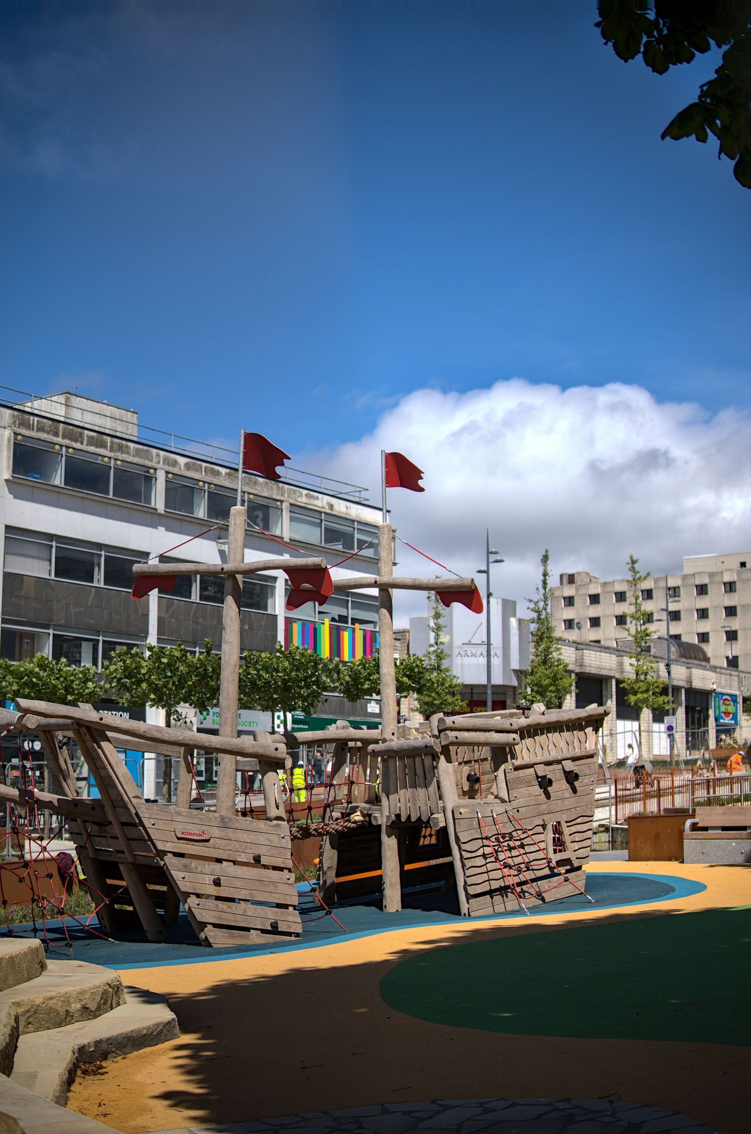 Wooden pirate ship playground structure with red flags in an urban park under blue sky.