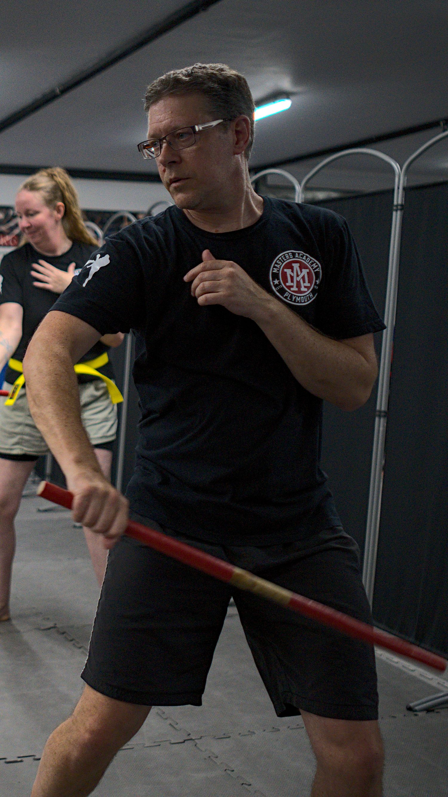 Male martial arts instructor in black uniform demonstrates a staff strike technique in a training facility.