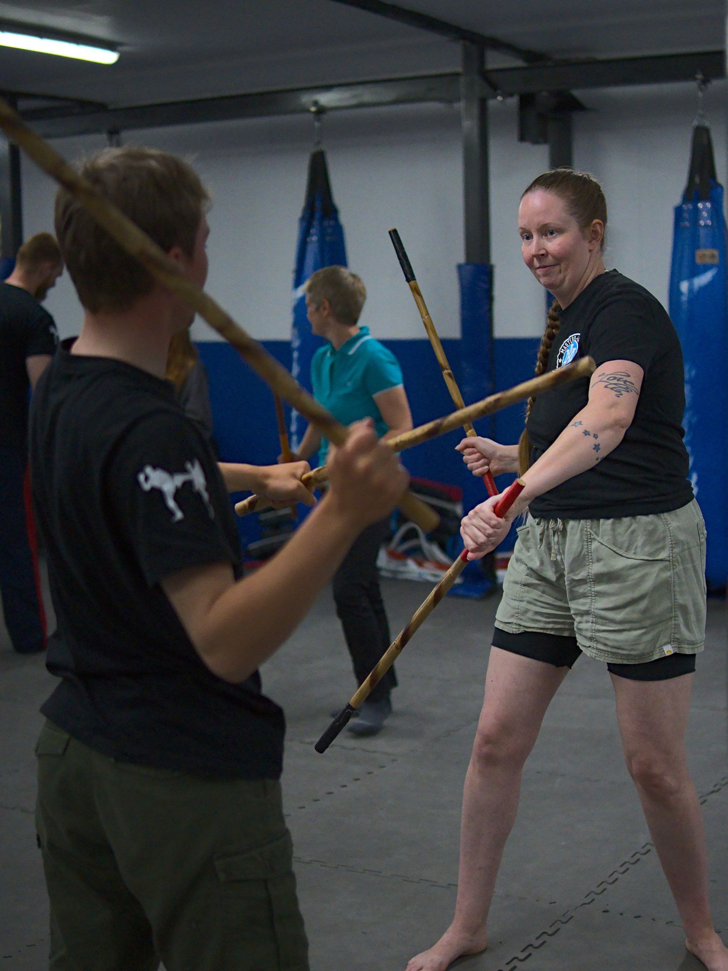 Two female martial artists smile while sparring with wooden sticks during an Arnis training session in a gym.