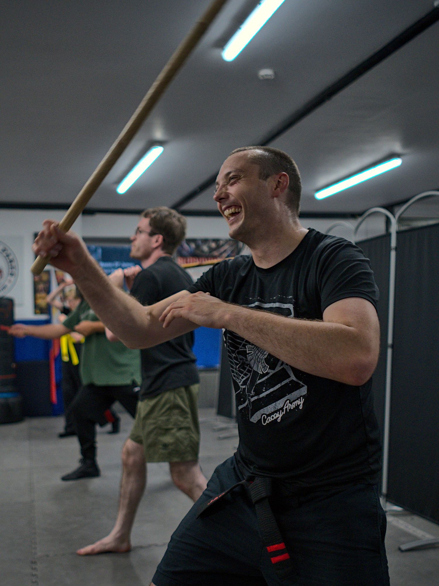 A smiling man practices stick fighting techniques with a wooden staff in a martial arts class.