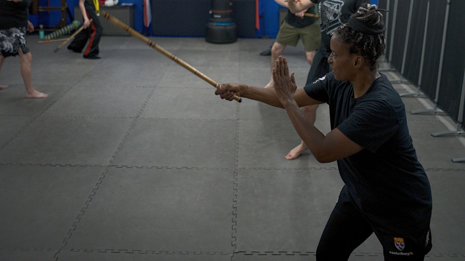 A martial artist practices stick fighting techniques with a wooden staff on a gym mat floor.
