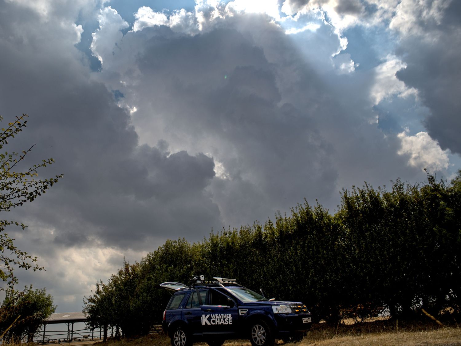 UK Weather Chase vehicle parked under dramatic storm clouds with dark skies and sunbeams breaking through.