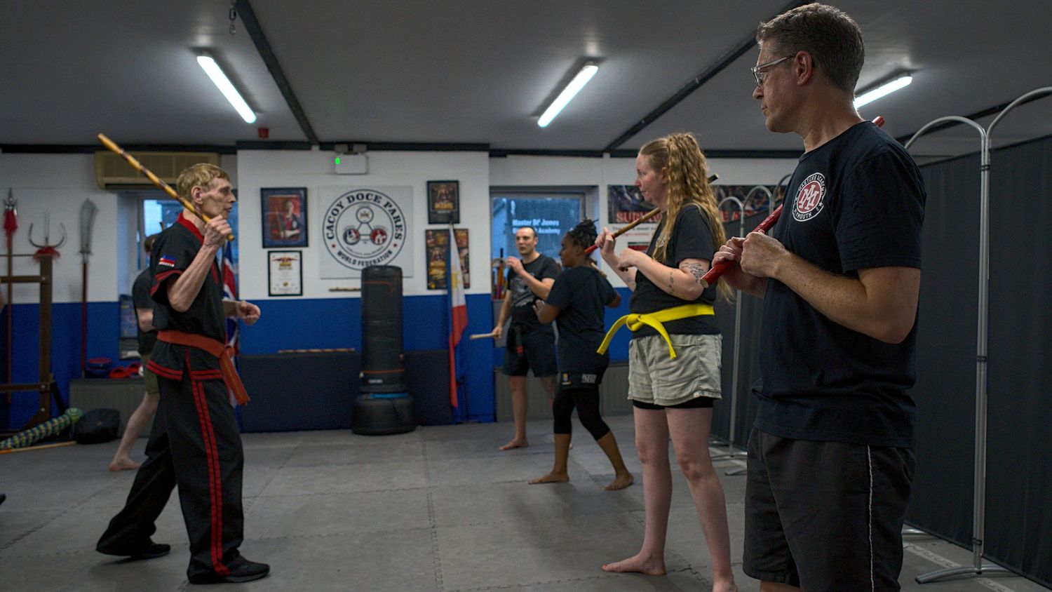 Martial arts instructor demonstrates stick fighting techniques to students of varying belt levels in a dojo.