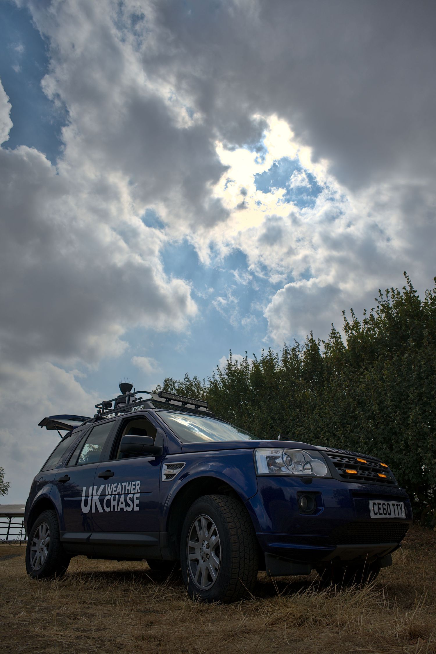 UK Weather Chase blue Land Rover parked in field beneath dramatic cloudy sky with sun breaking through clouds.