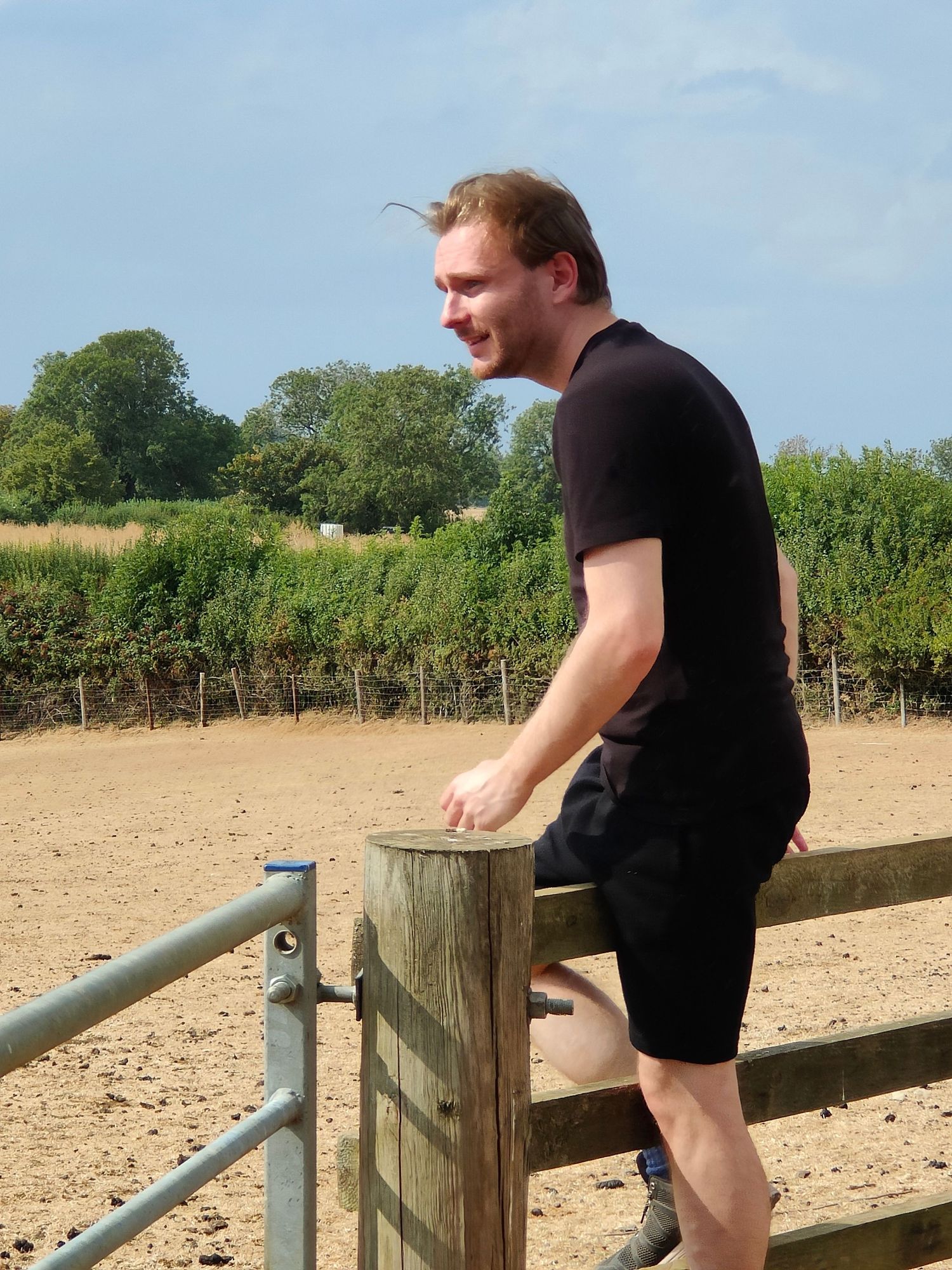Young man in black t-shirt and shorts leaning on a wooden farm gate in a sunny rural English countryside.