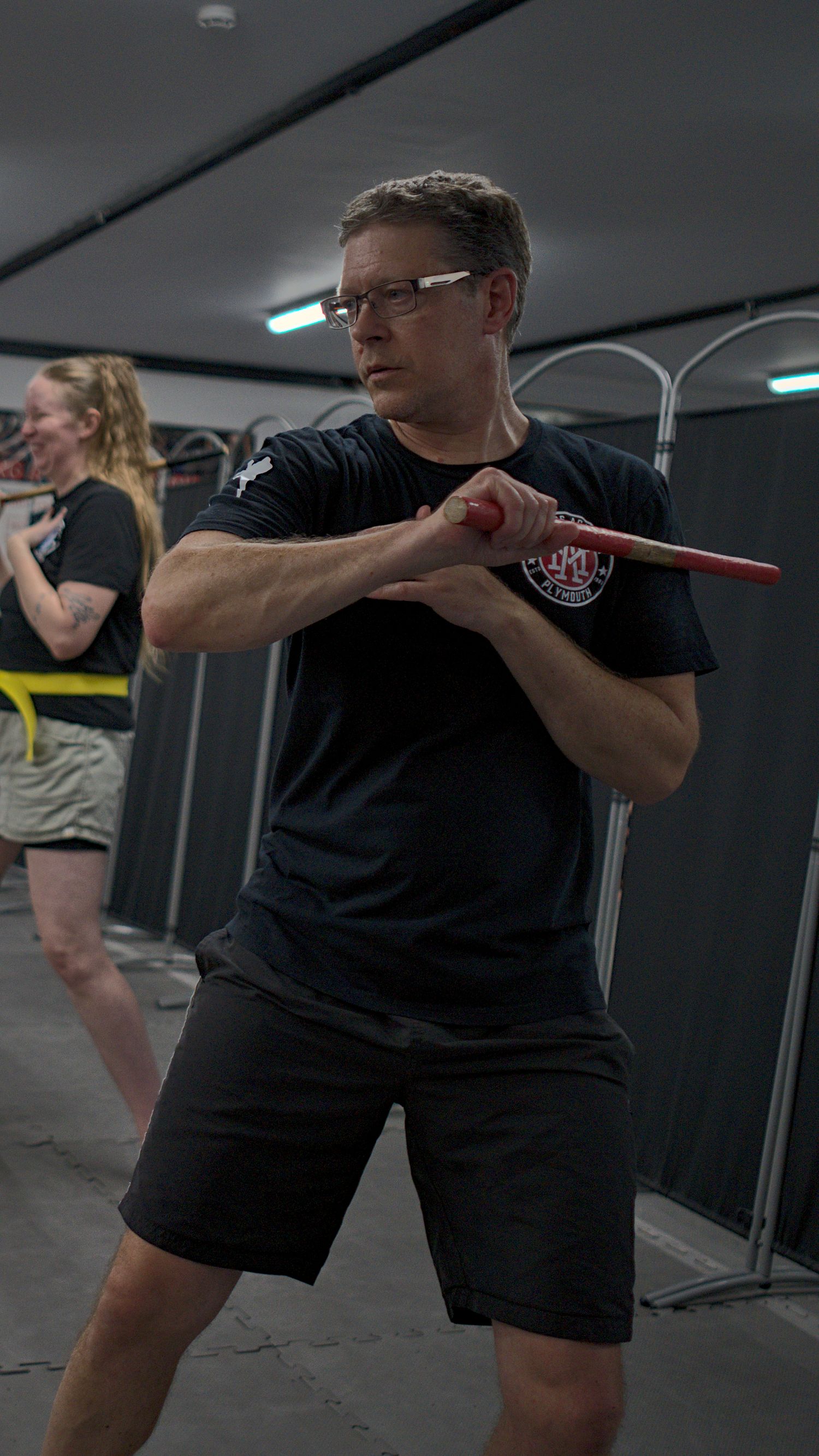 Male martial arts practitioner in black uniform executes a defensive stick fighting position in a gym.