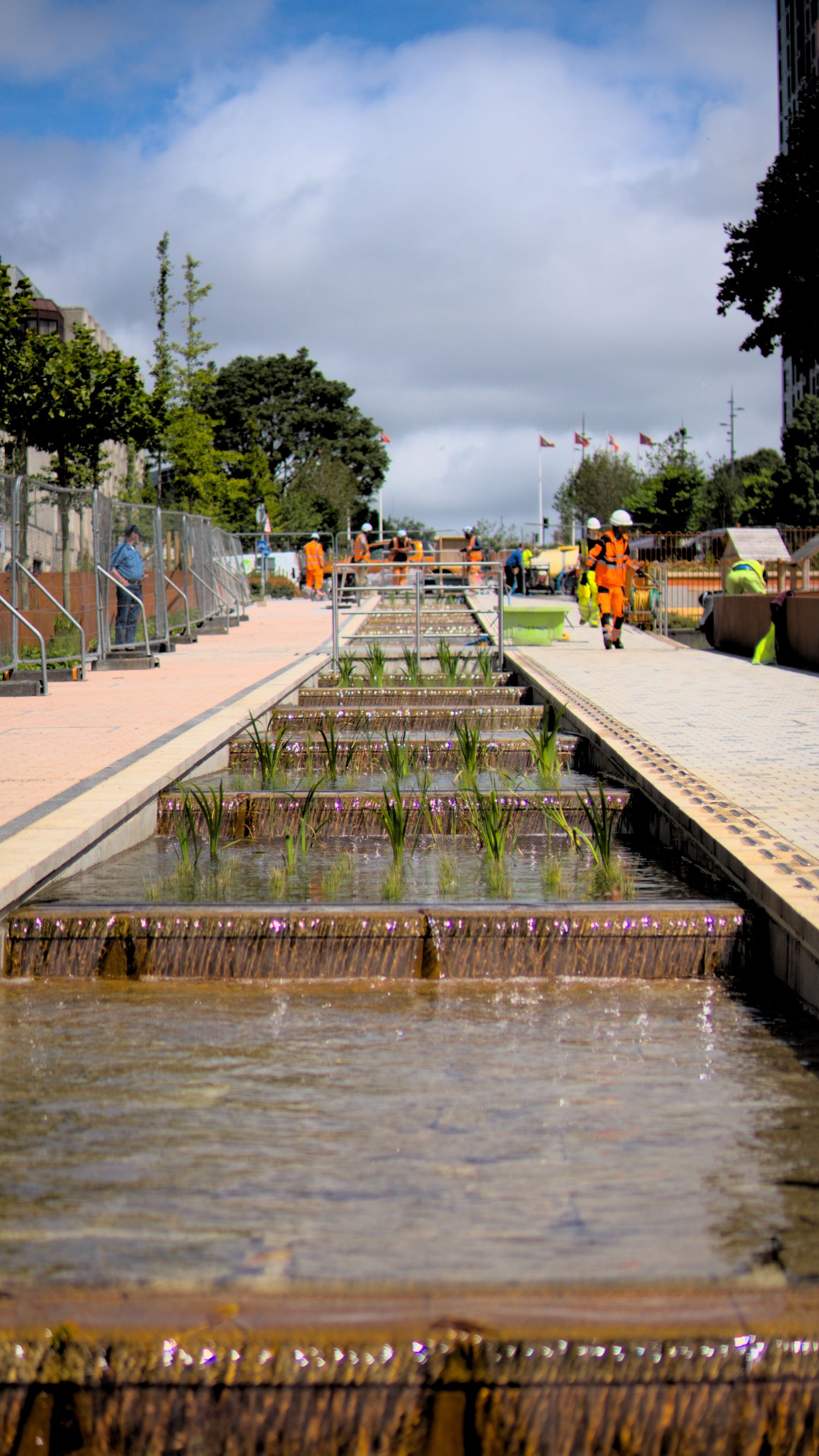 Construction workers in orange safety gear near a stepped water feature fountain in an urban park.