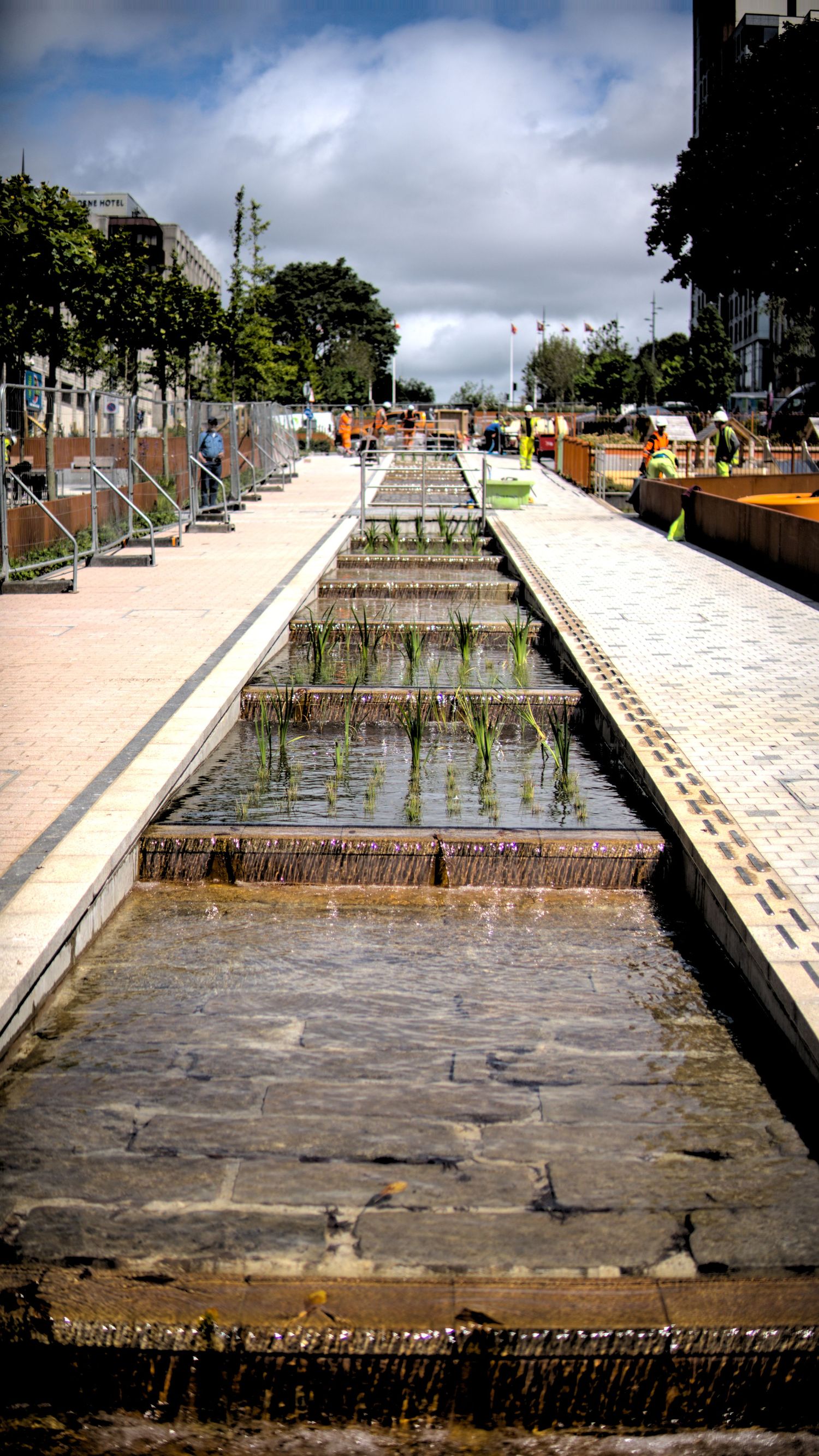 Stepped cascading water feature with aquatic plants in an urban plaza under a cloudy sky.