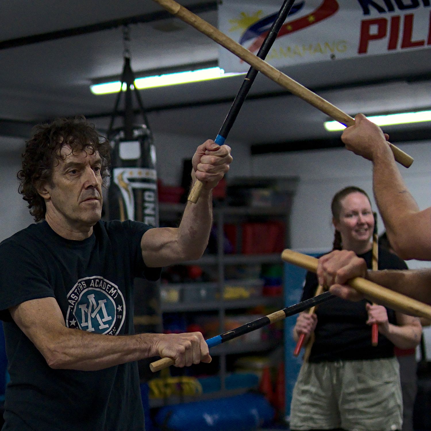 Students practice Filipino stick fighting Arnis techniques in a martial arts gym with punching bags in background.