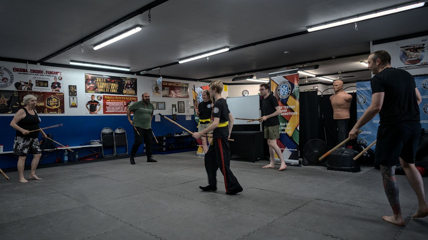 Instructor leads a group stick fighting martial arts class in a well-equipped indoor gym.