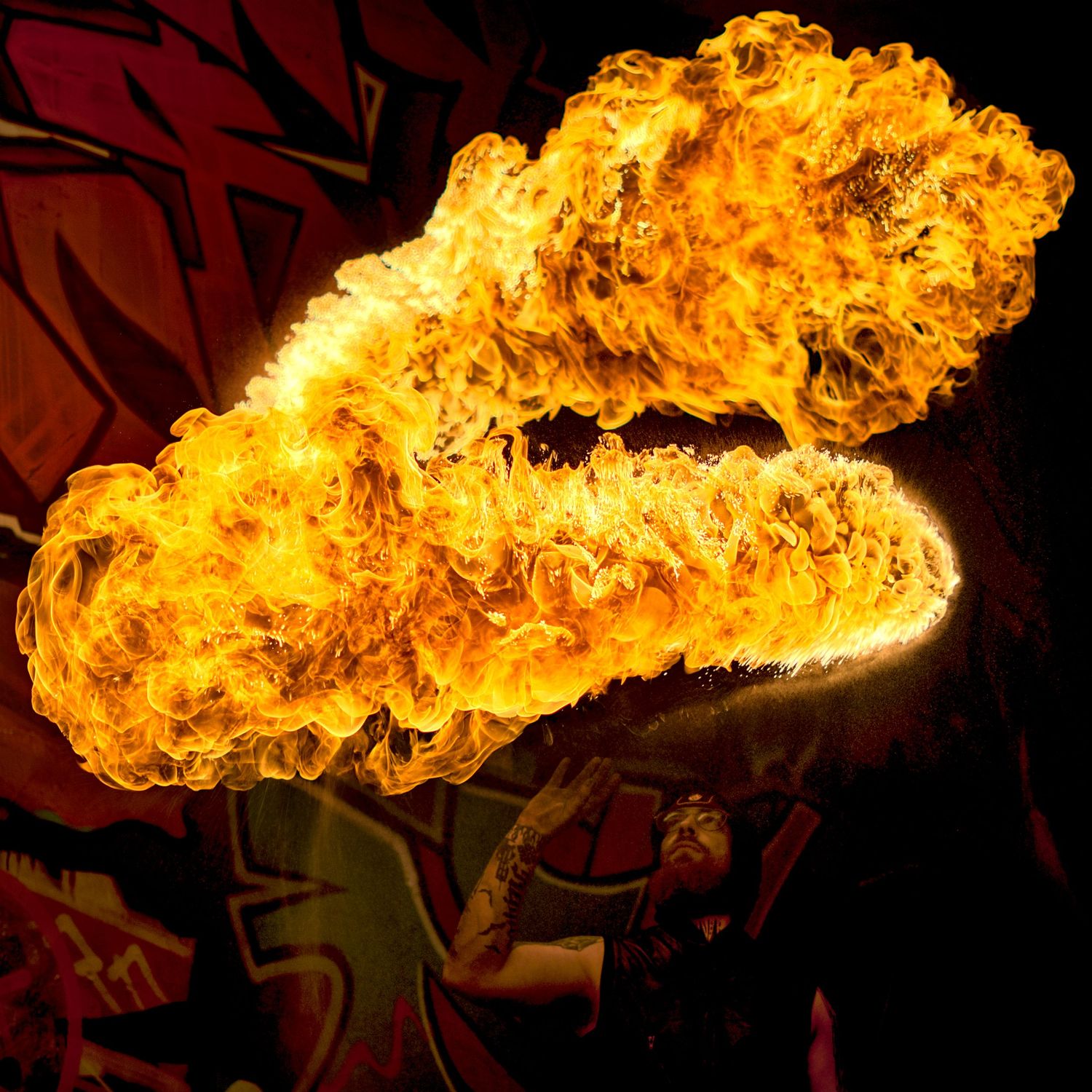Fire breather exhaling a massive ball of flame against a dark graffiti backdrop.