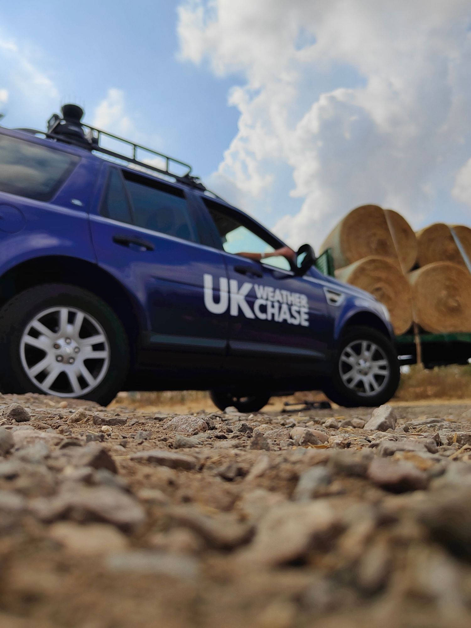 UK Weather Chase blue Land Rover parked near hay bales in dry field under partly cloudy sky.