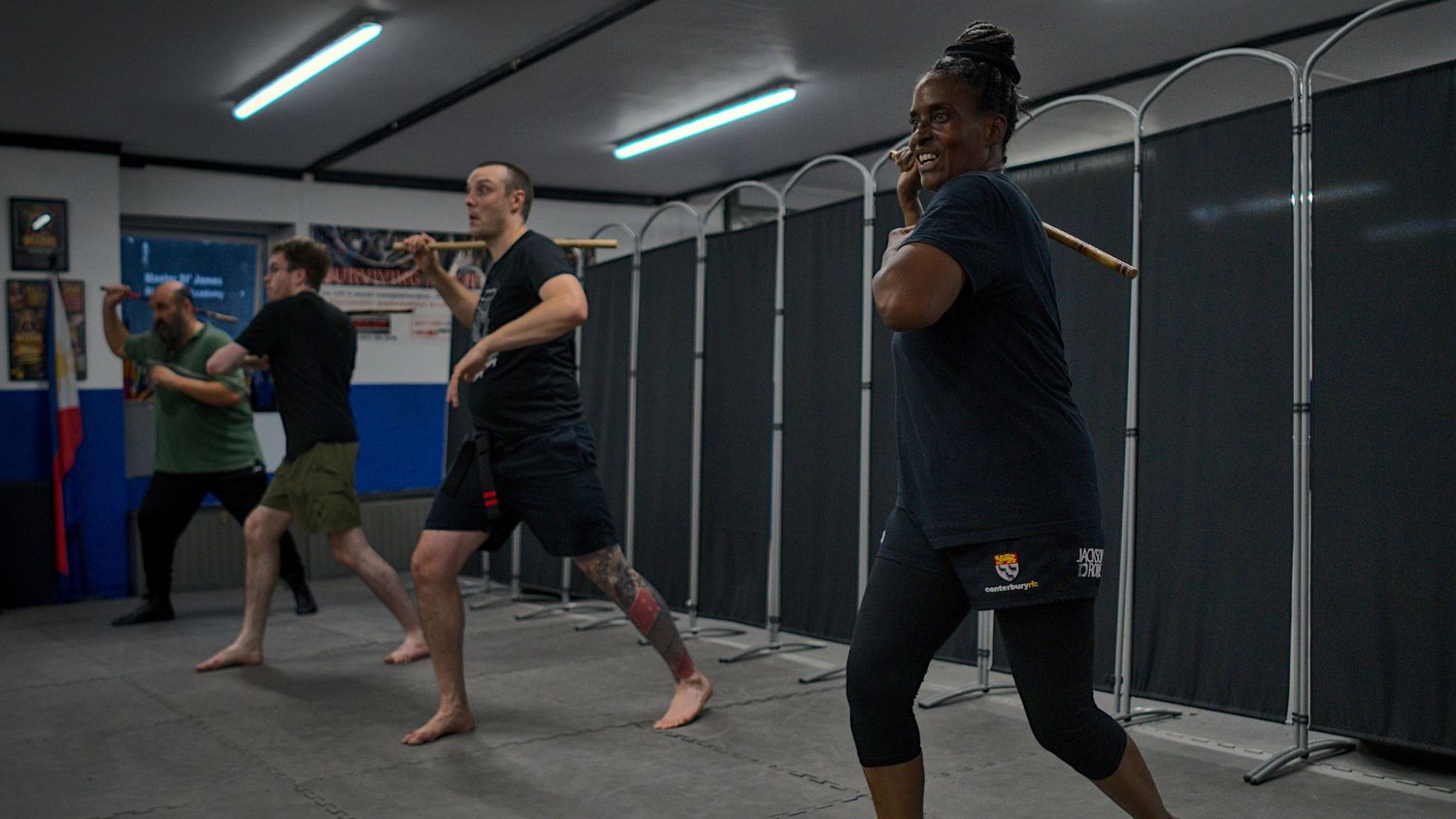 Group of martial arts students practice stick fighting drills in an indoor training gym.
