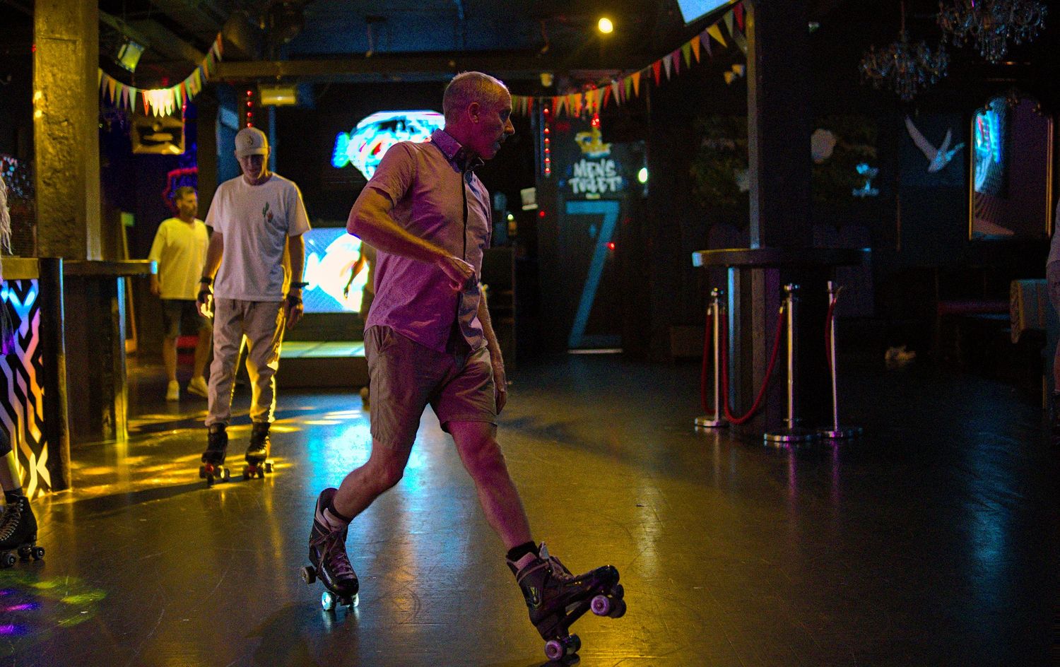Bald man roller skating in neon-lit indoor skating rink at night.