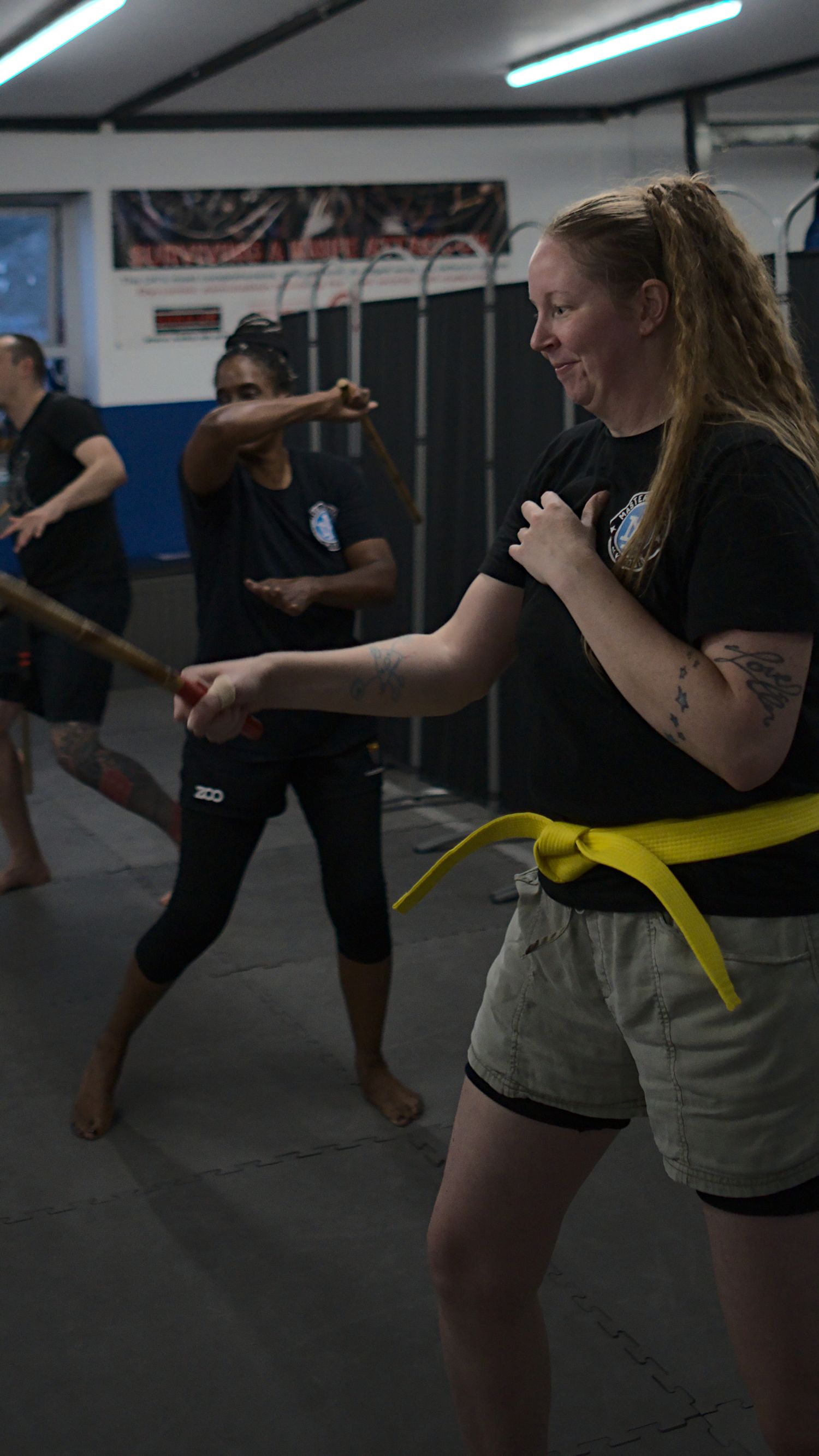 Smiling female yellow belt martial arts student practices stick fighting techniques in a group class.