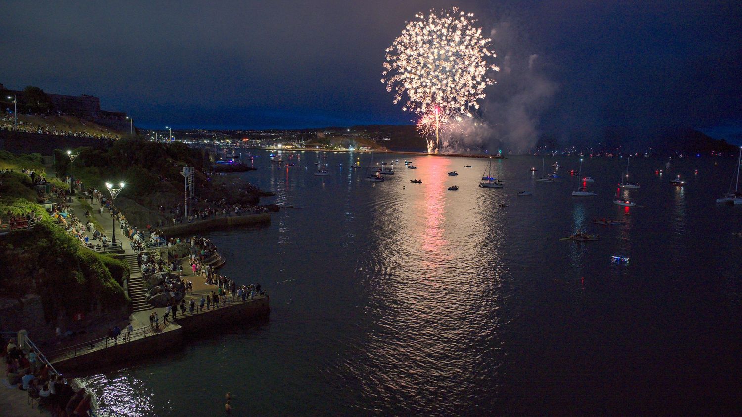 Colorful fireworks explode over a harbor at night, reflecting on water surrounded by boats and illuminated waterfront buildings.