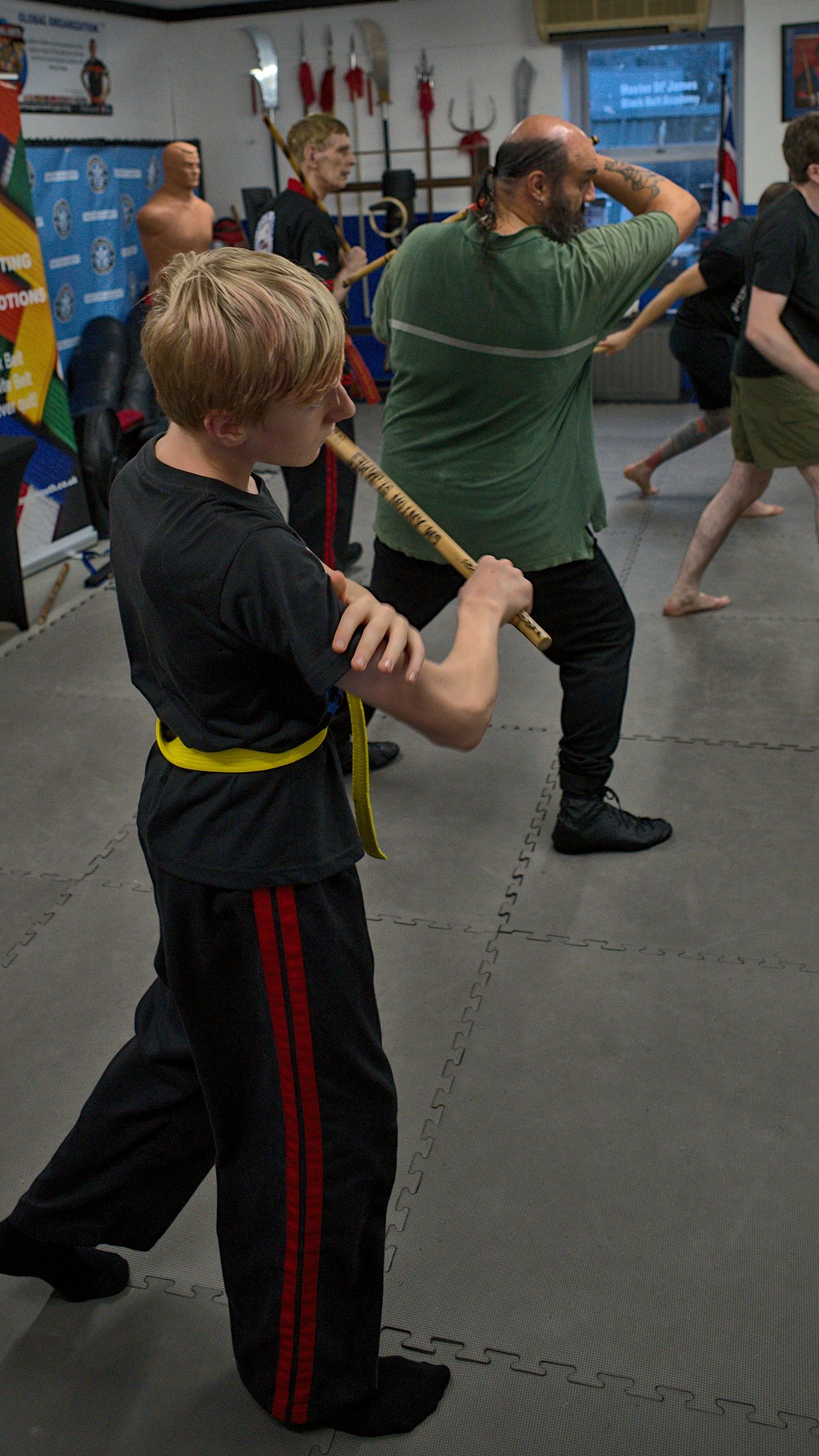 Young martial arts student with yellow belt trains with a wooden staff under instructor supervision in a dojo.