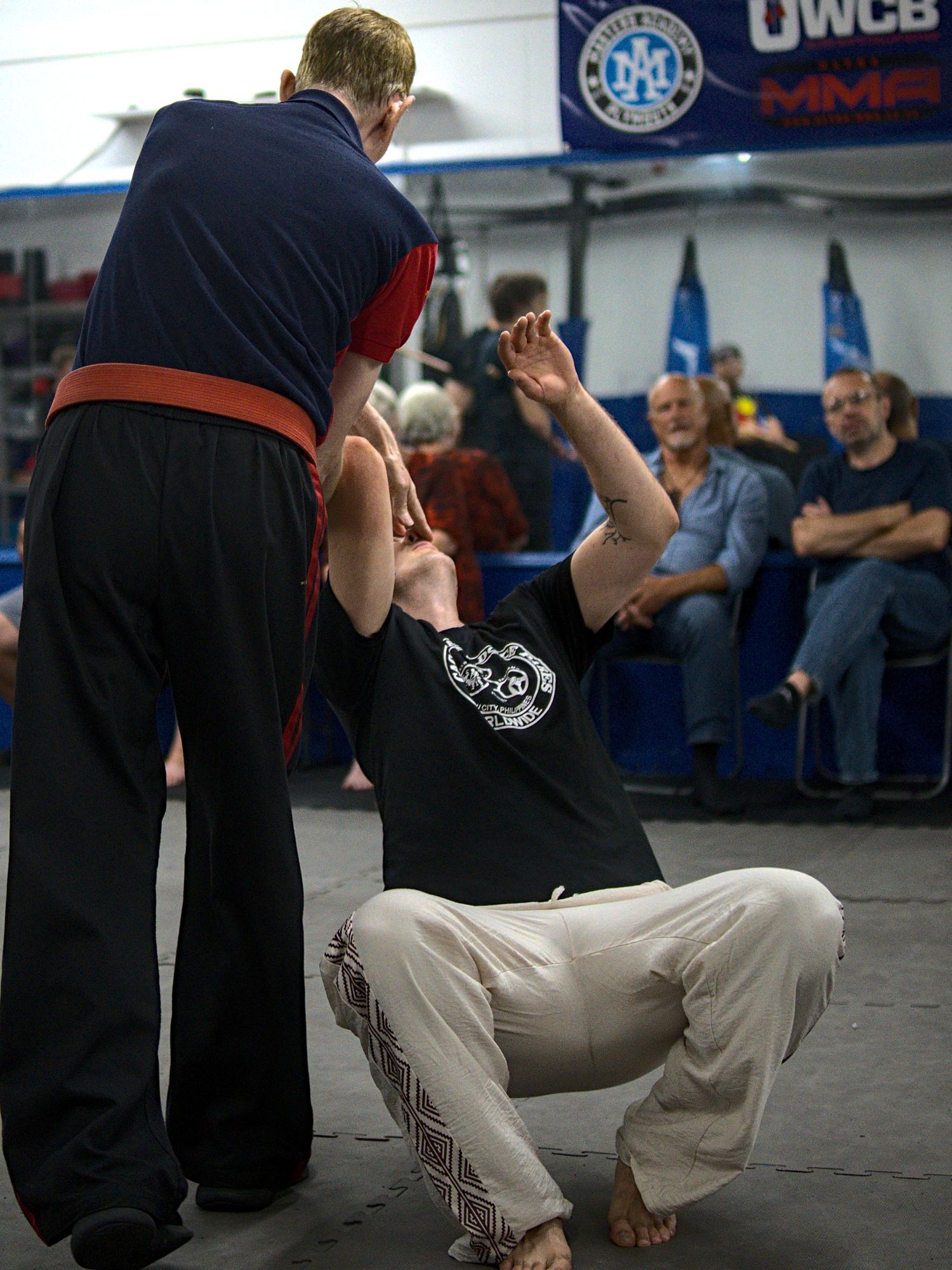 Martial arts instructor guides kneeling student through ground defense technique during class at indoor gym.