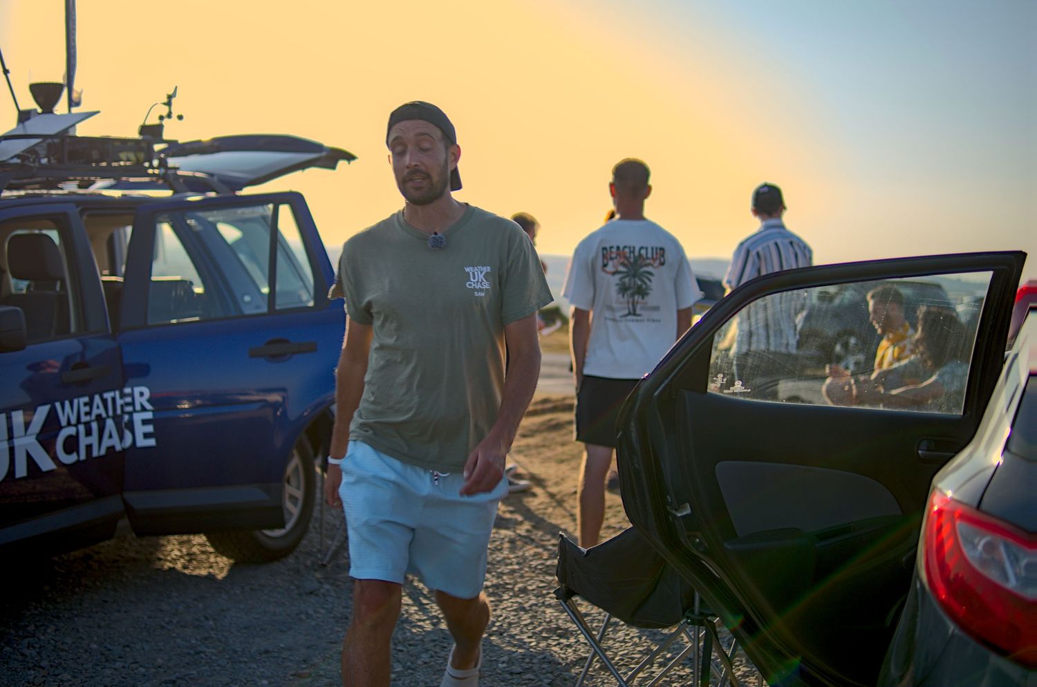 Man standing beside UK Weather Chase vehicle at sunset with group of people in background.