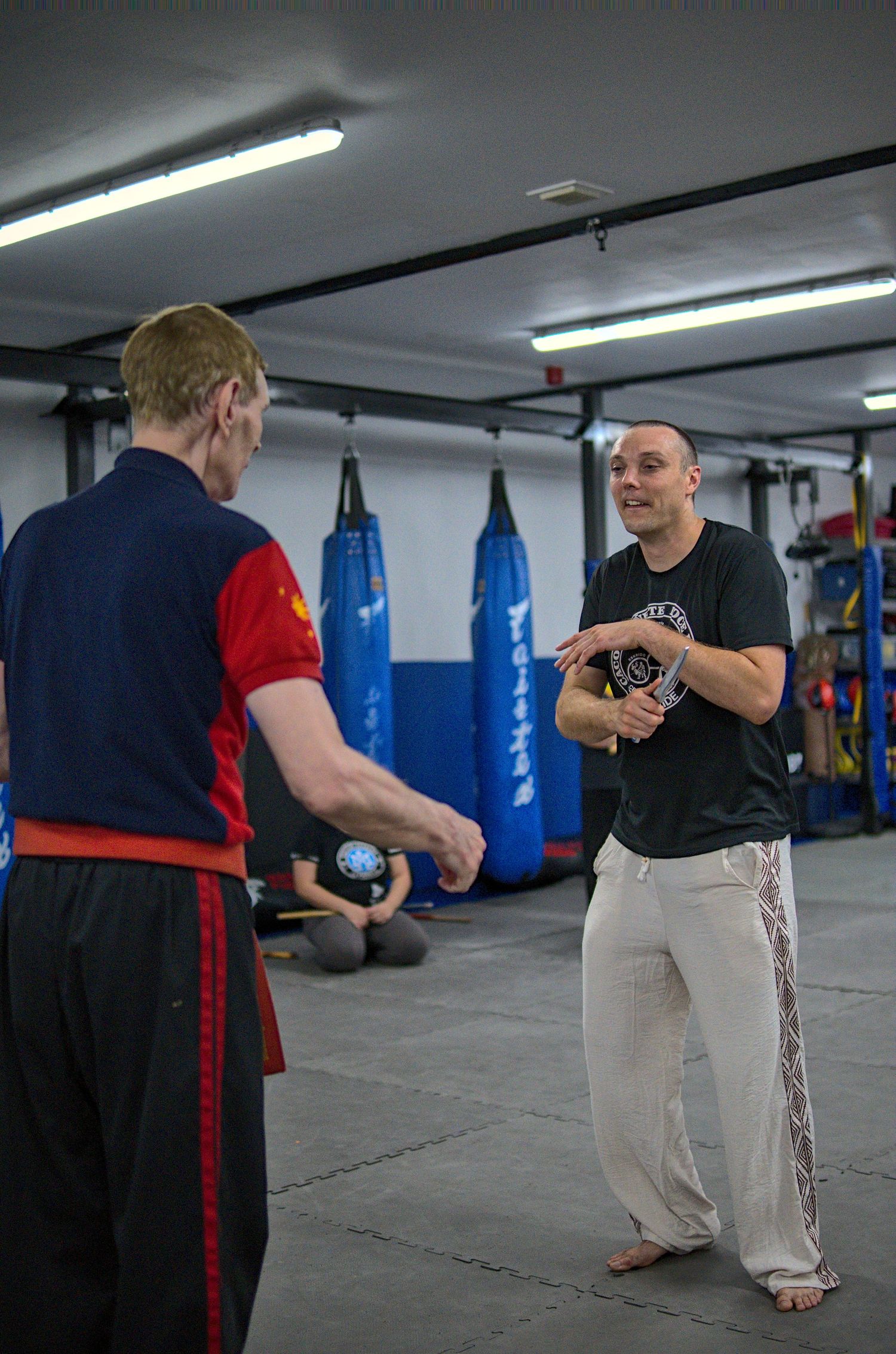 Two martial artists interact during a training session near blue punching bags.
