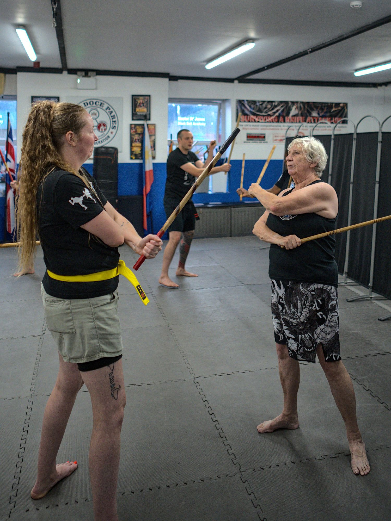 Two women practice stick fighting techniques with wooden staffs during martial arts class at indoor dojo.