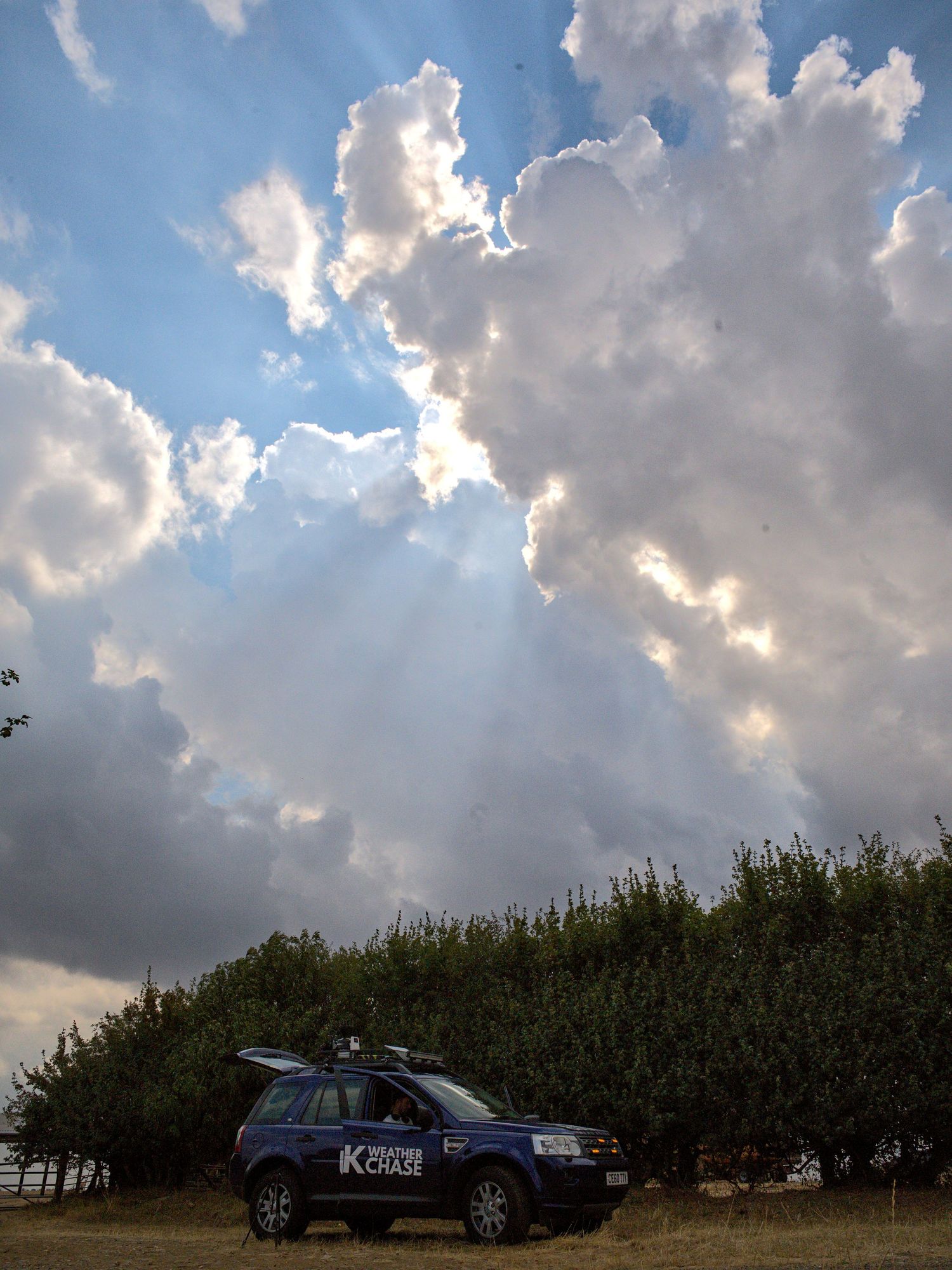 UK Weather Chase car parked beside hedgerow under dramatic clouds with sunbeams breaking through.