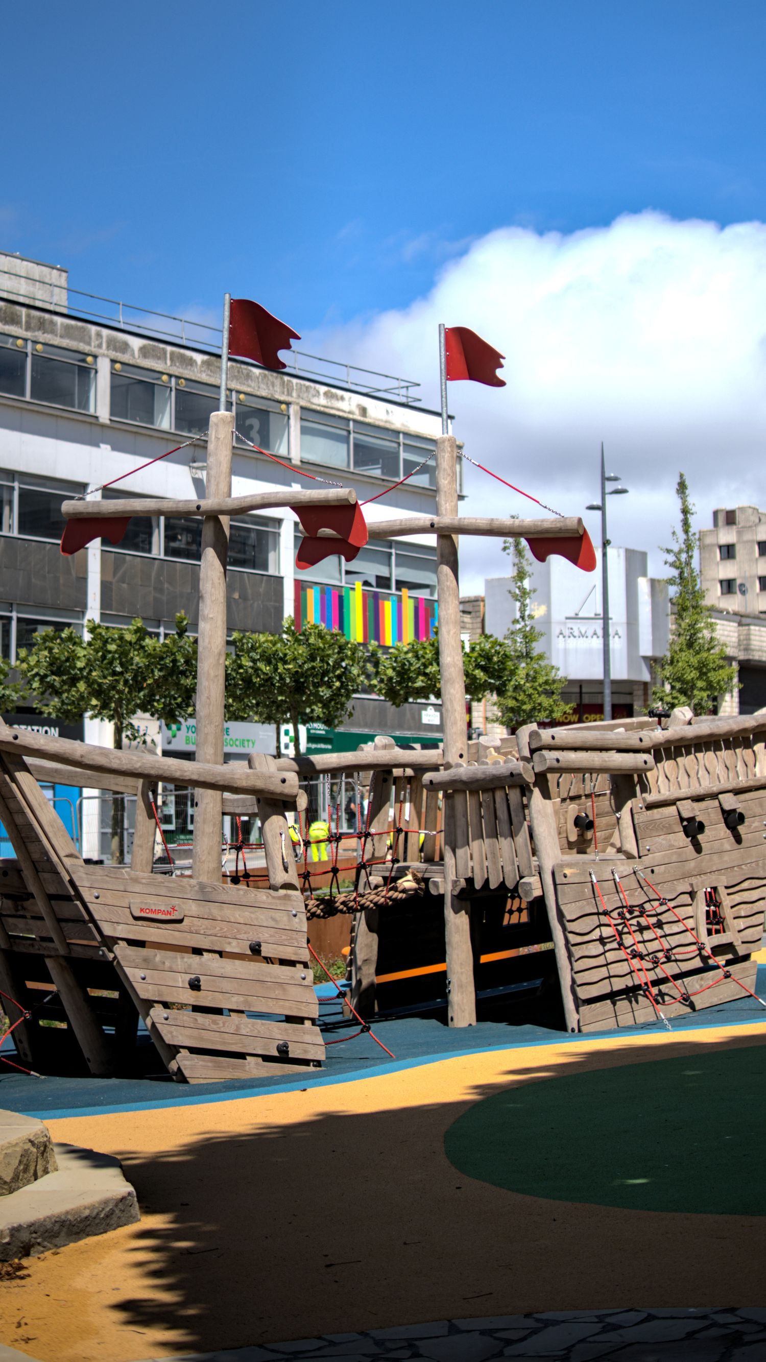 Wooden pirate ship playground with red flags and climbing ropes in a city park.