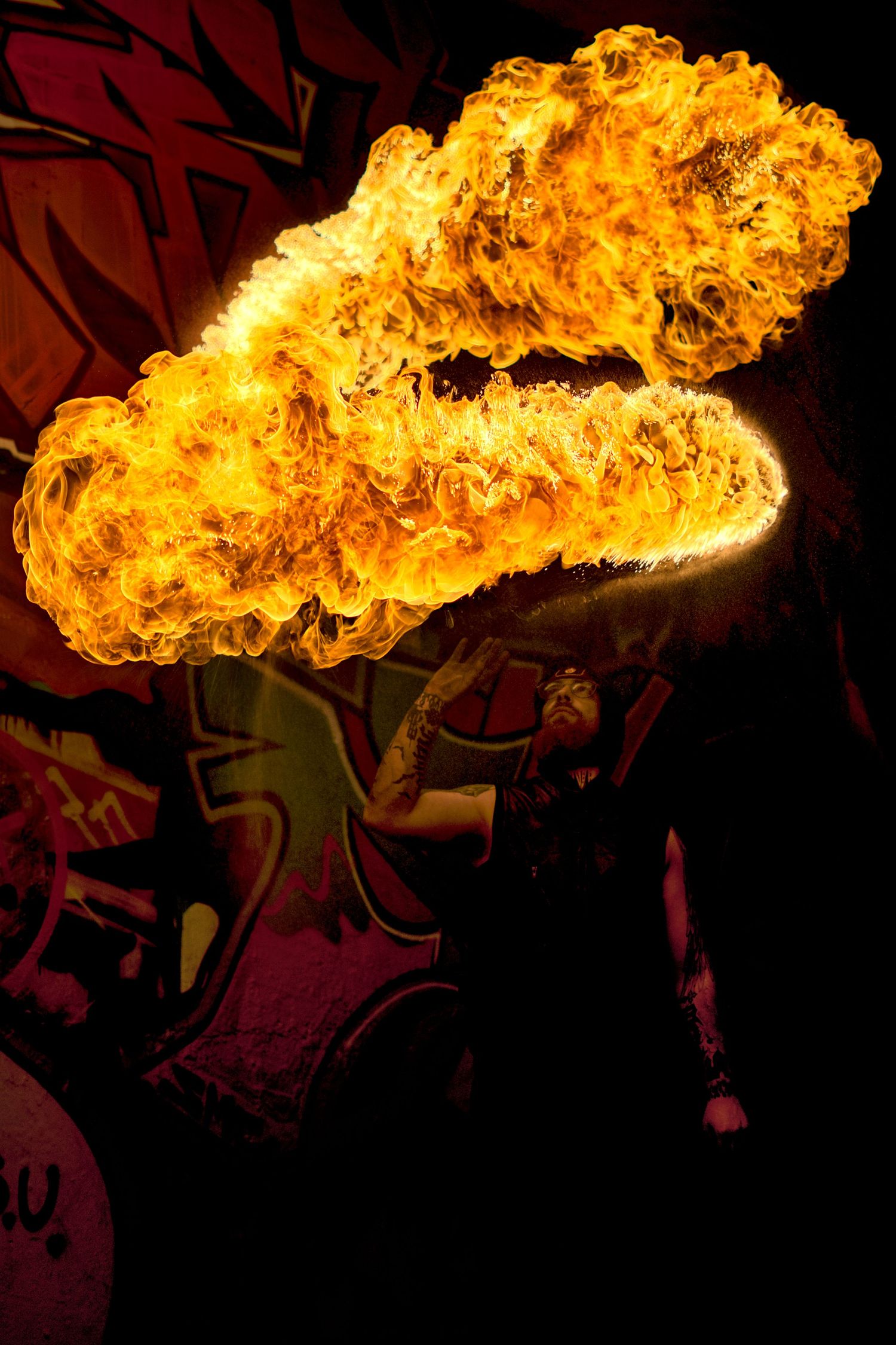 Fire breather producing a large horizontal fireball against a colorful graffiti wall.