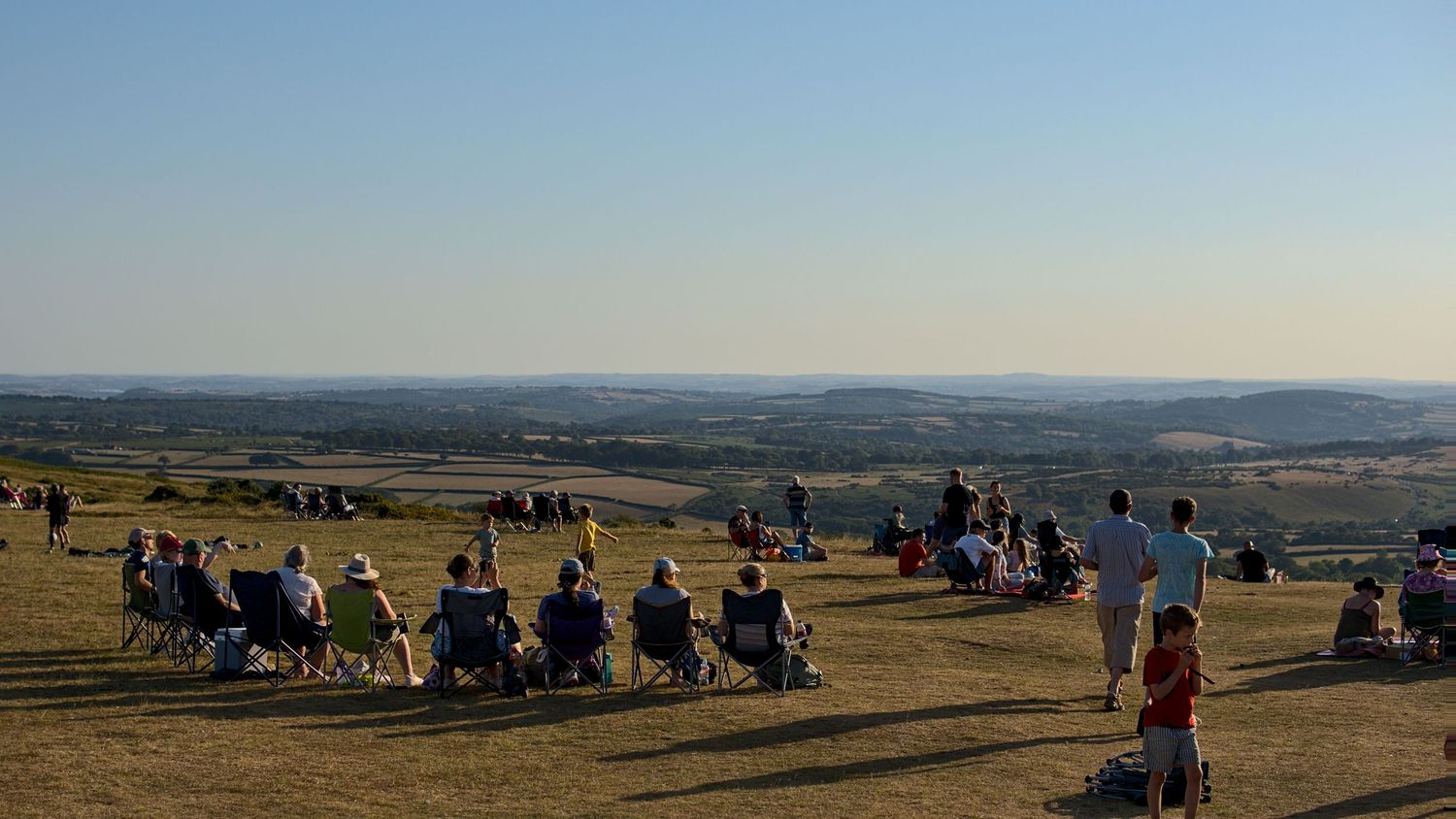 Large crowd of people sitting in chairs on hilltop enjoying panoramic countryside views on clear day.