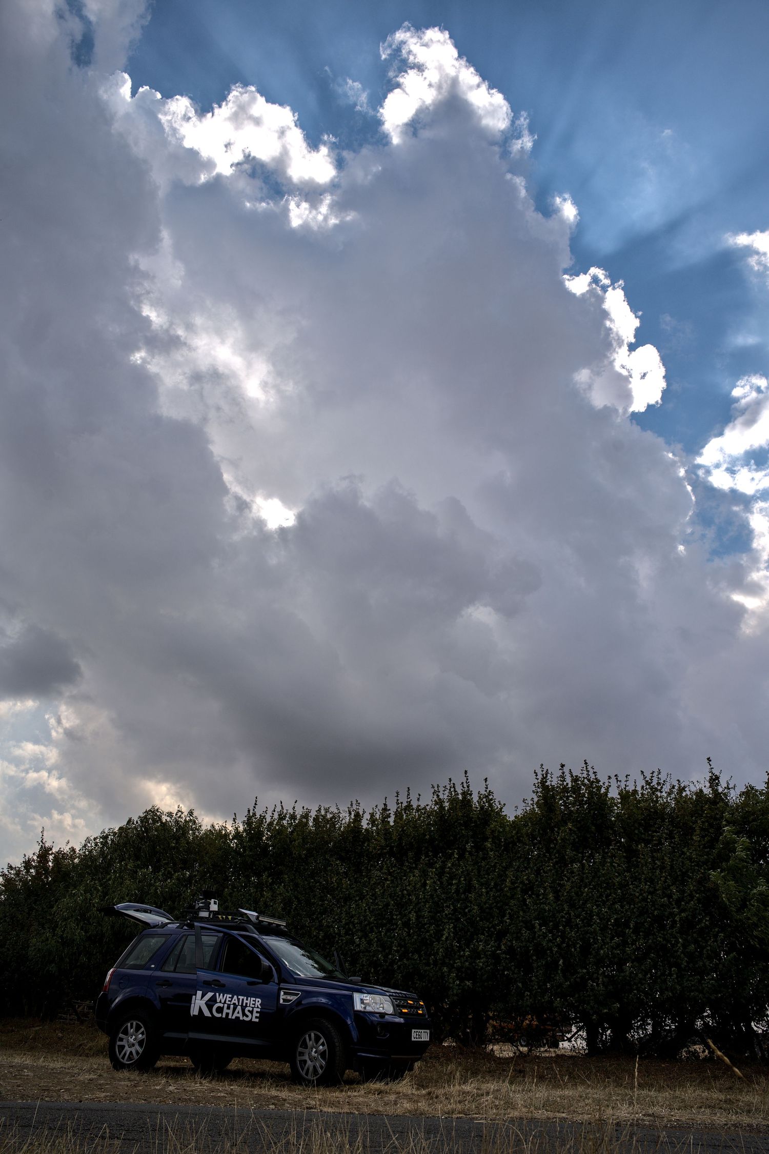 UK Weather Chase vehicle parked beside tall hedgerows beneath a massive dramatic cumulonimbus storm cloud.