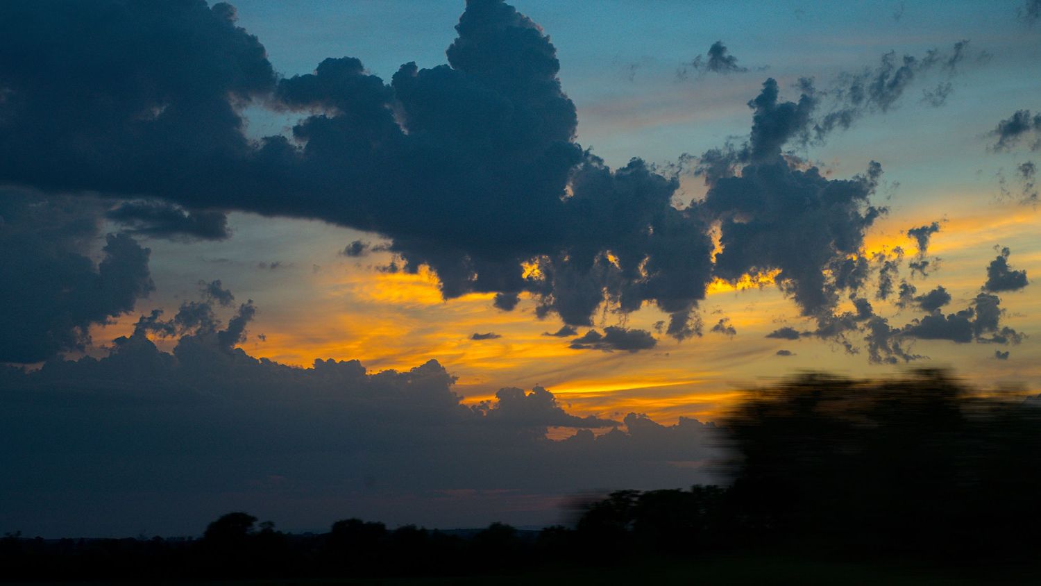 Dramatic sunset sky with dark storm clouds illuminated by vivid orange and yellow light over silhouetted trees.