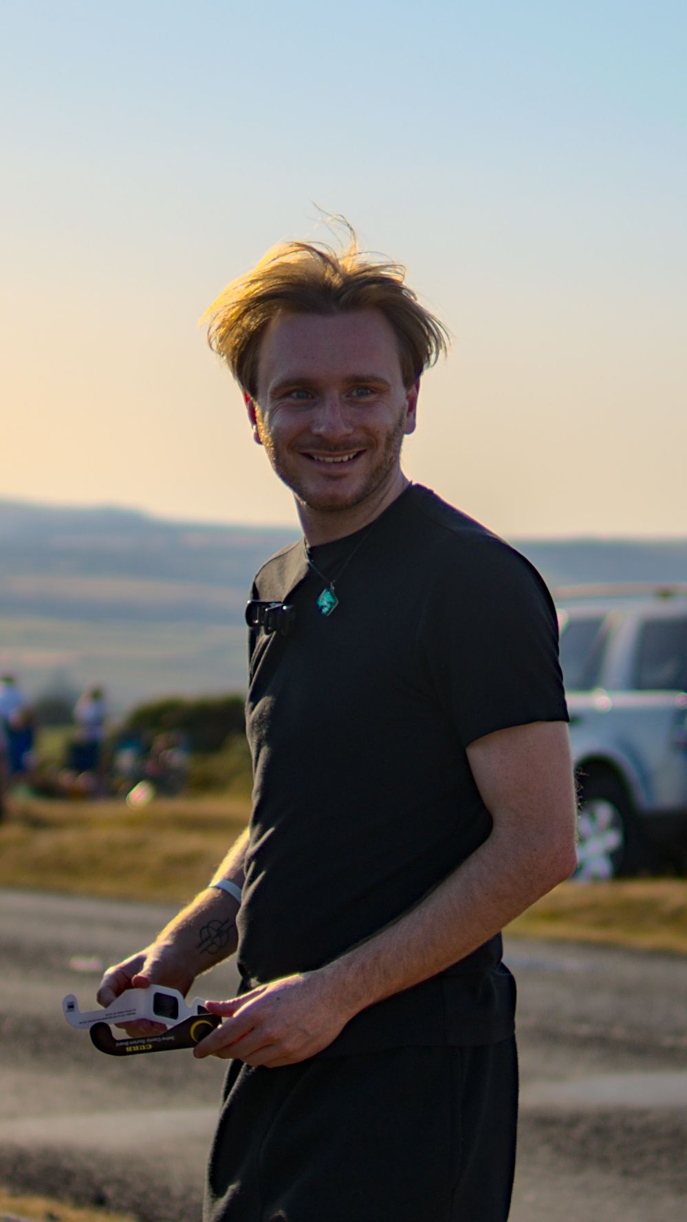 Smiling young man in black t-shirt holding camera equipment outdoors at golden hour sunset.