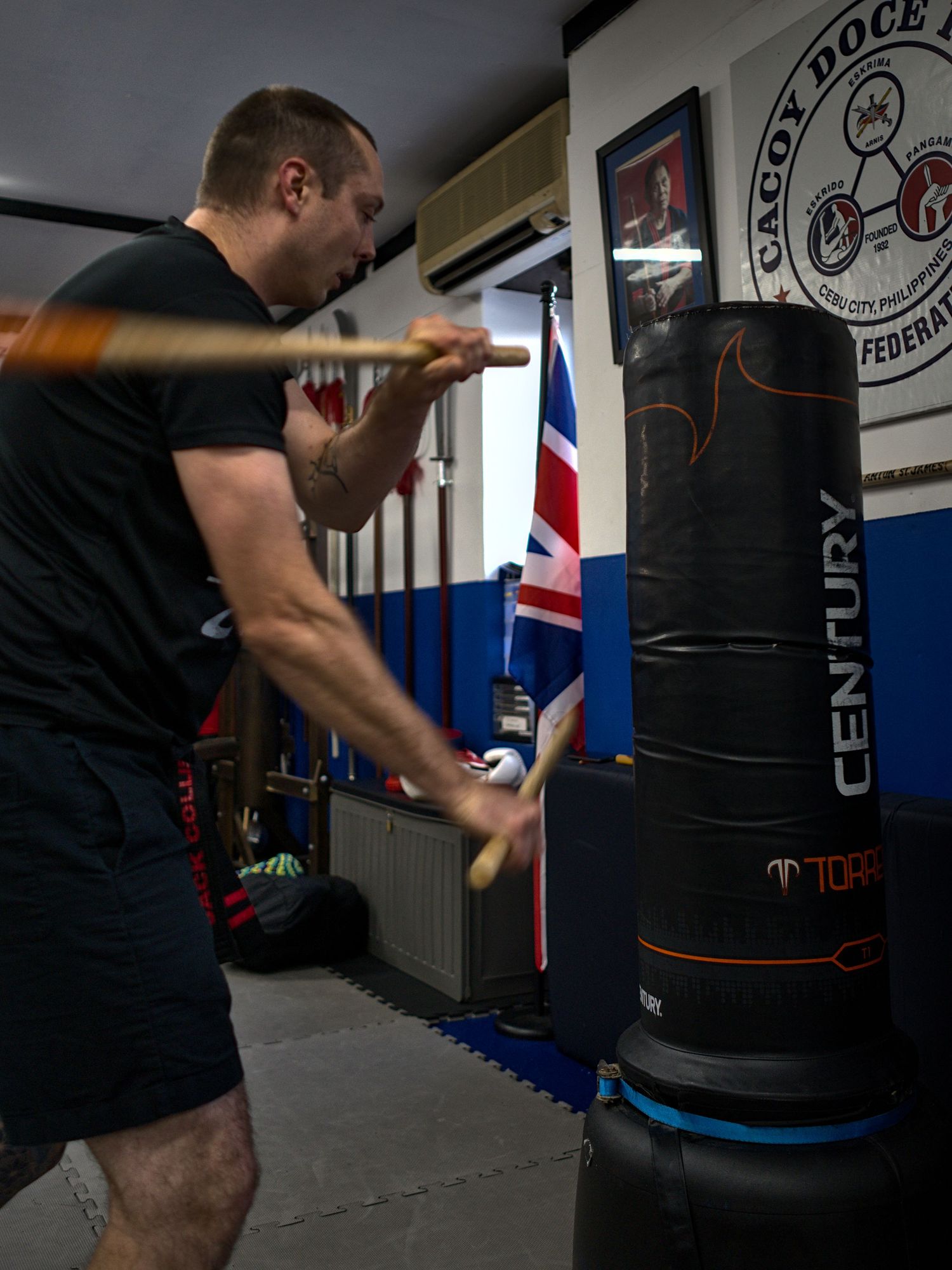 A martial artist strikes a Century Torso punching bag with wooden sticks in a gym.