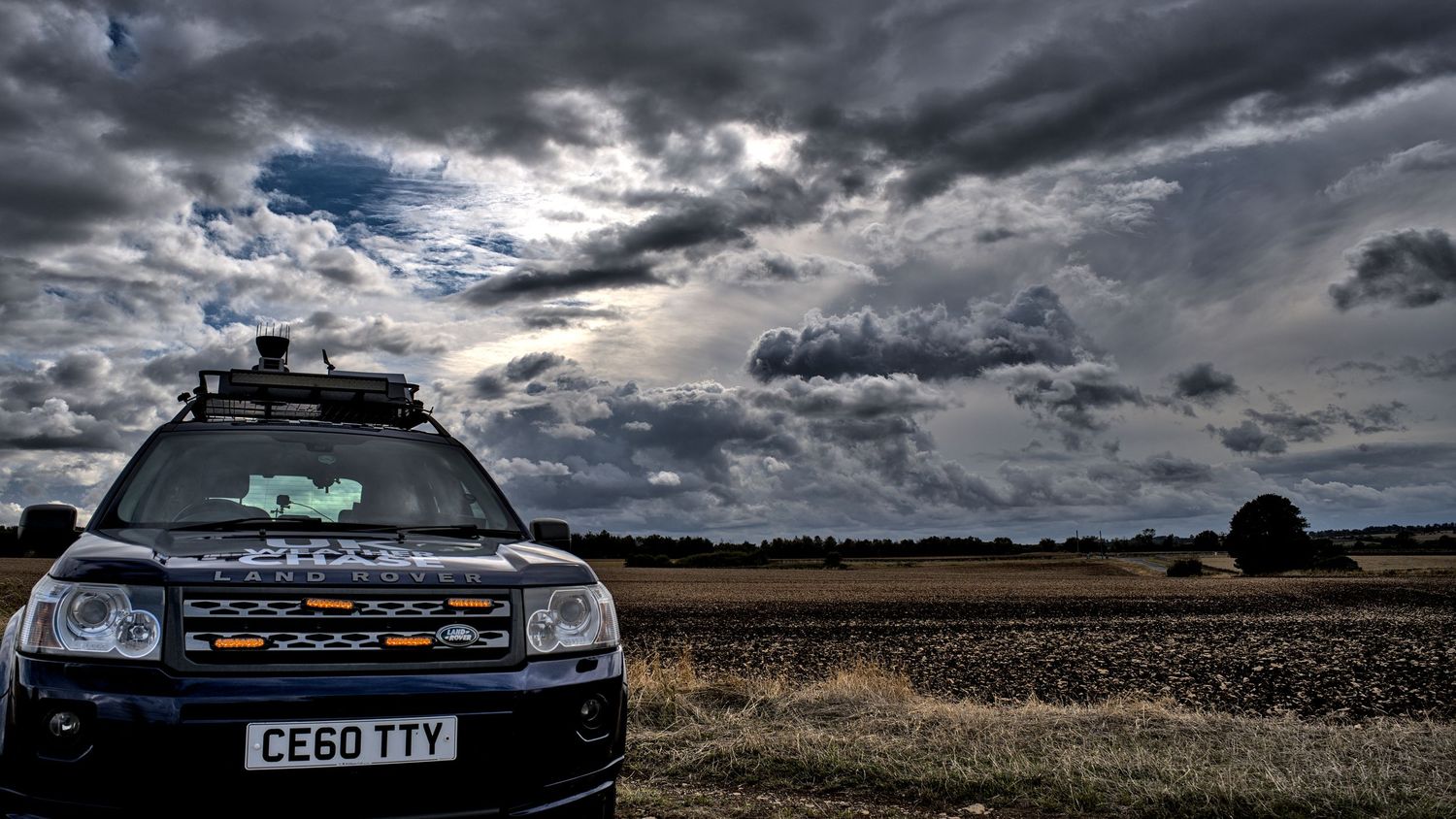 A UK Weather Chase Land Rover CE60 TTY sits in a rural field beneath a moody overcast sky.