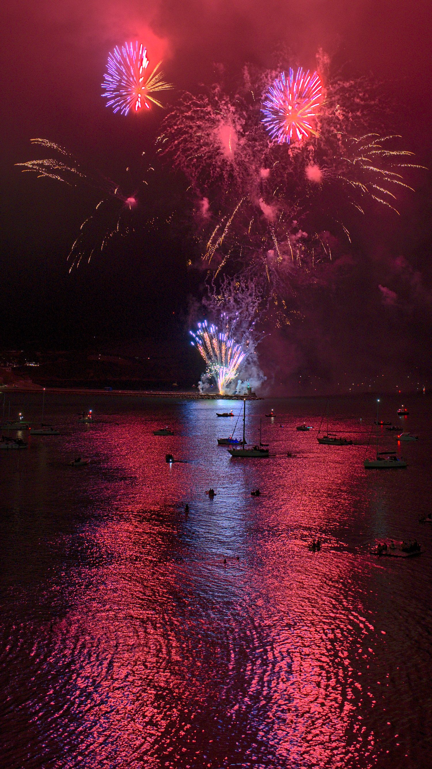 Vibrant fireworks display over a harbor at night, with colorful reflections shimmering on the water and boats silhouetted below.