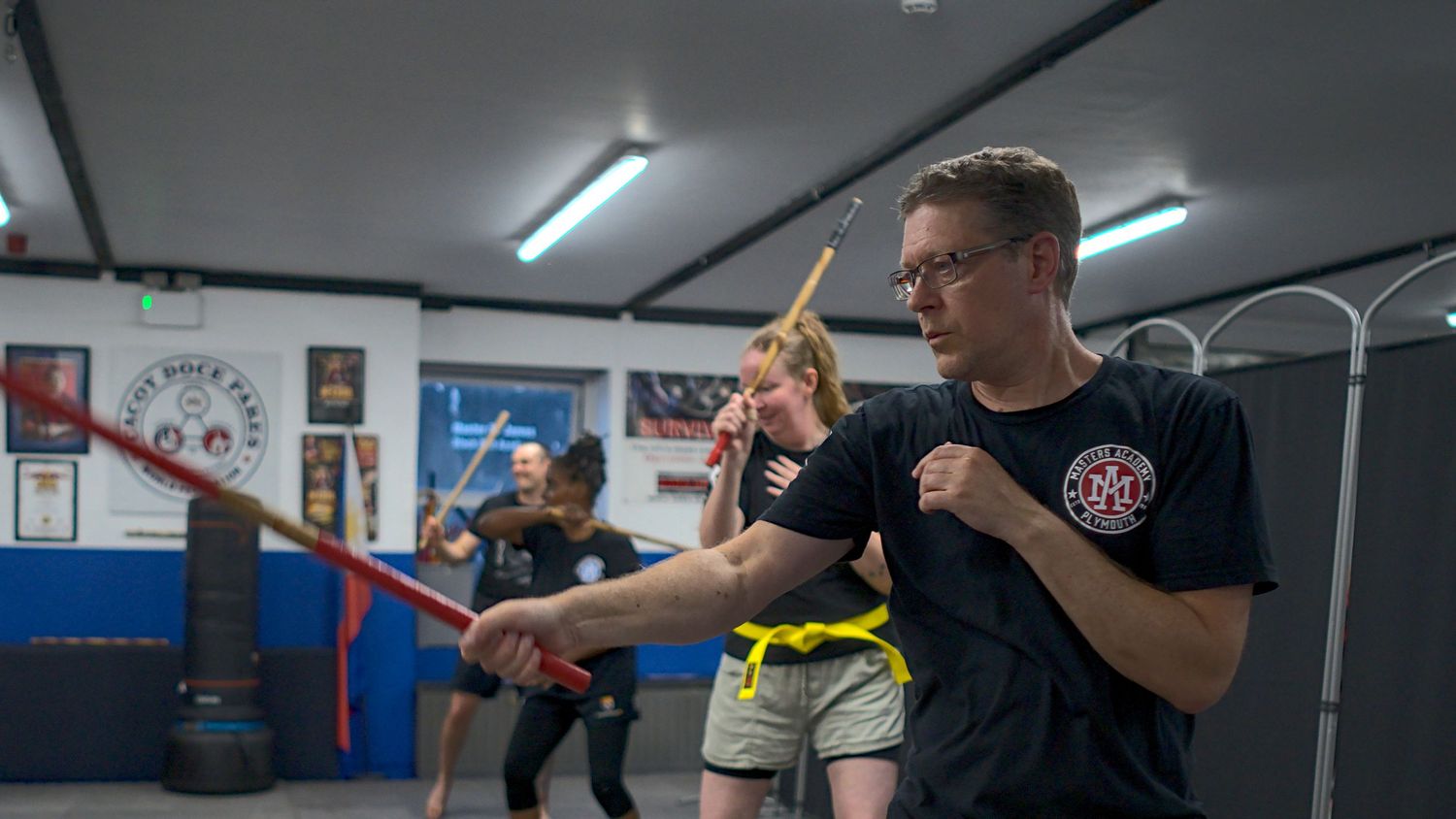 Two martial arts students spar with training sticks during a weapons class in an indoor dojo.