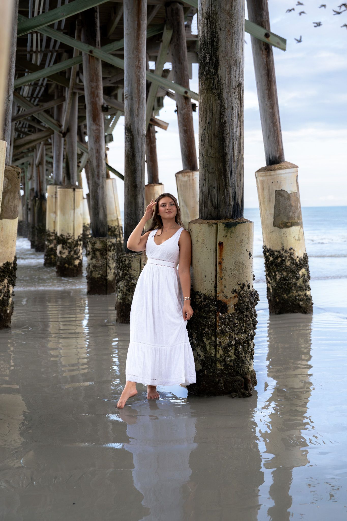 Woman in white maxi dress standing barefoot under a beach pier with ocean waves in the background.