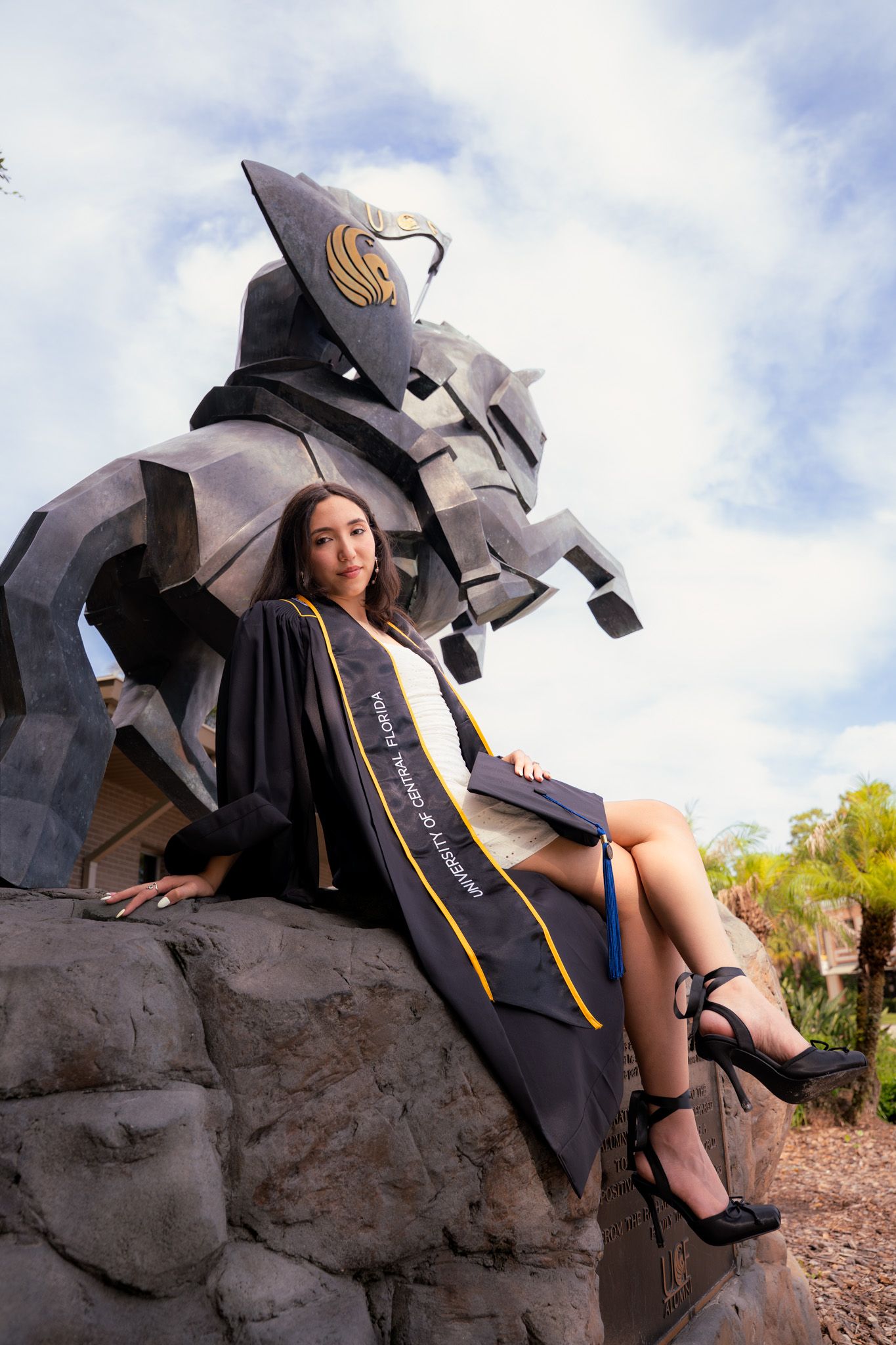 Female graduate in black gown sits on university mascot statue against cloudy sky.