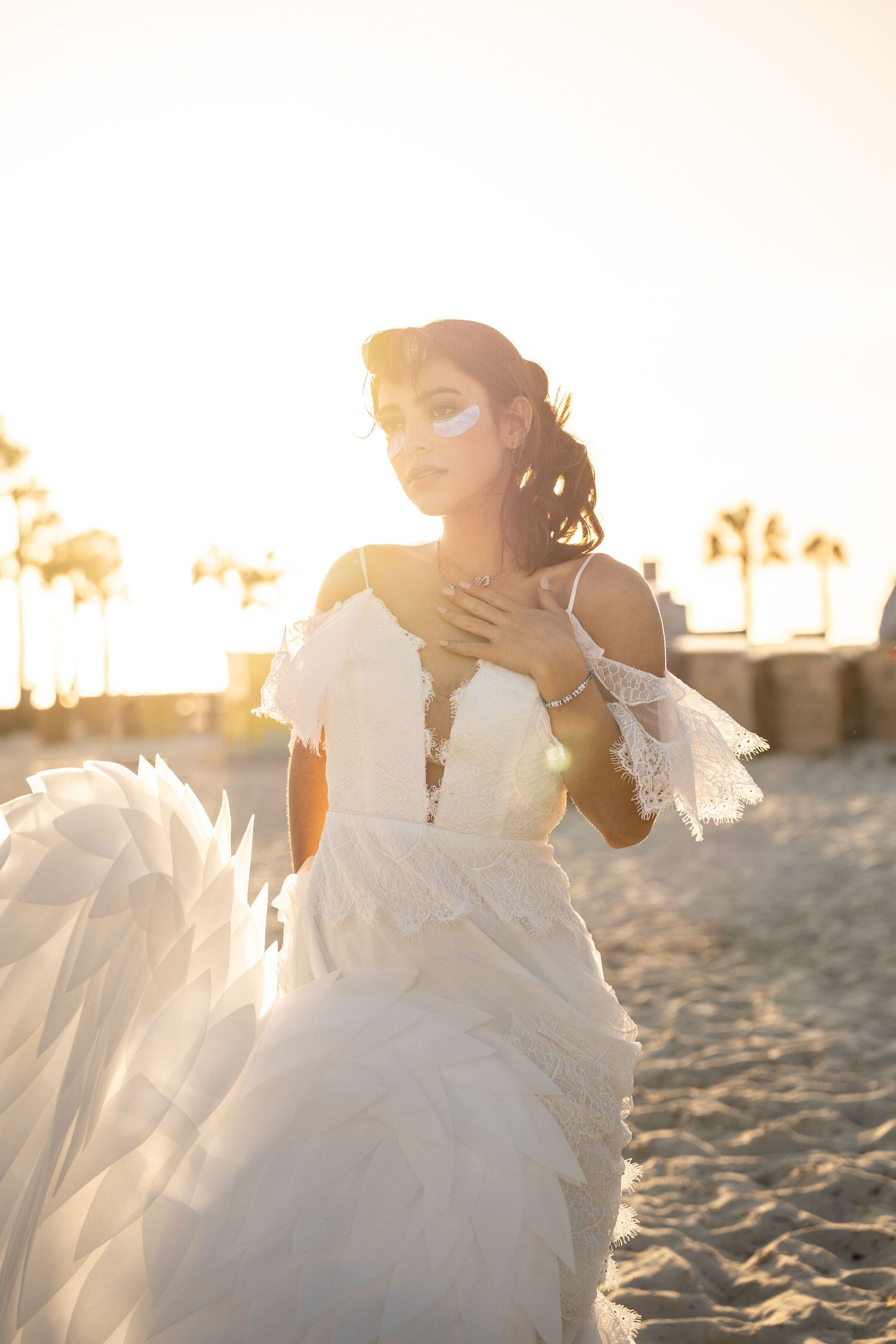 Bride in white wedding gown poses on sandy beach at golden sunset with palm trees in background.