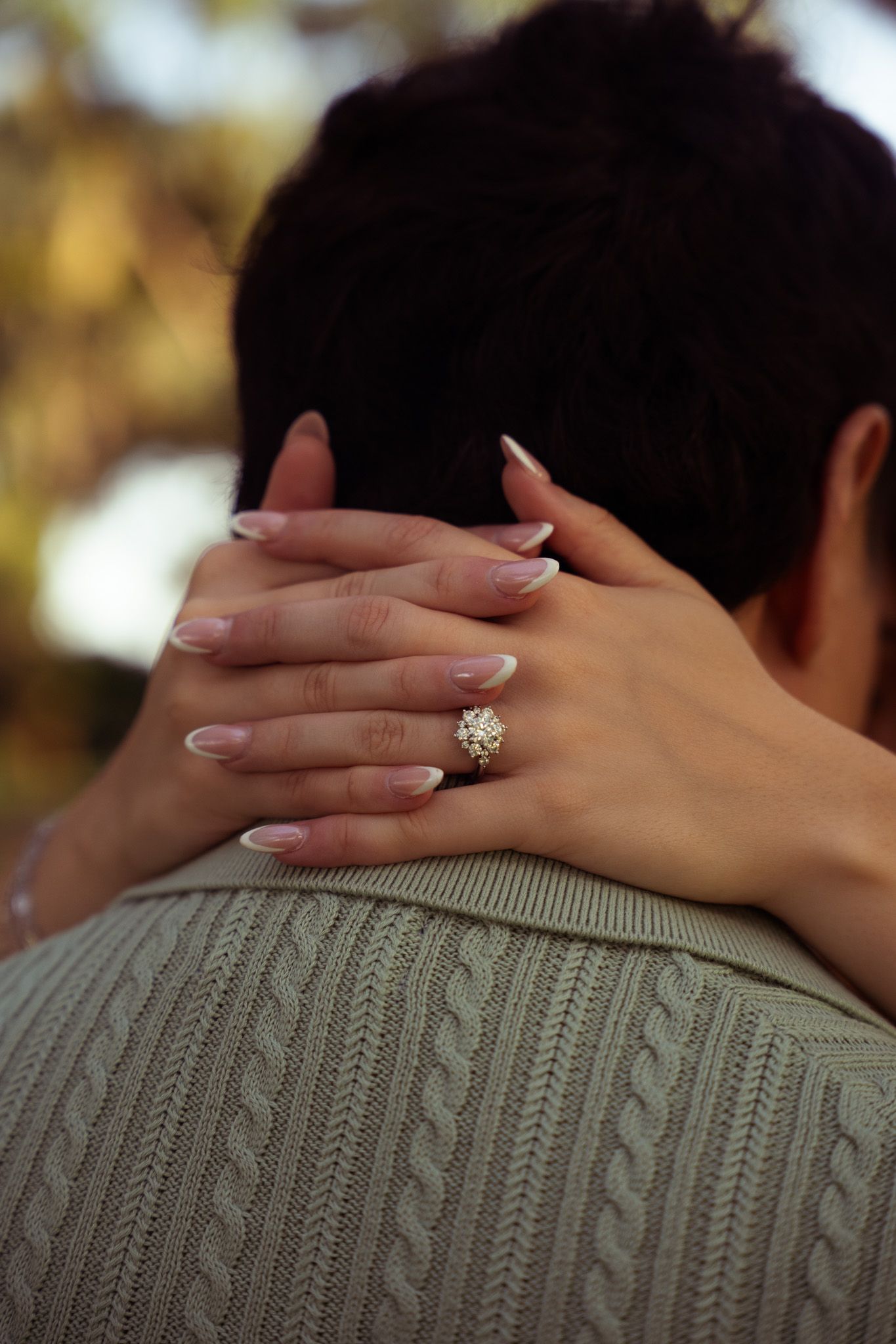 Close-up of woman's hand with a floral diamond engagement ring resting on man's shoulder.