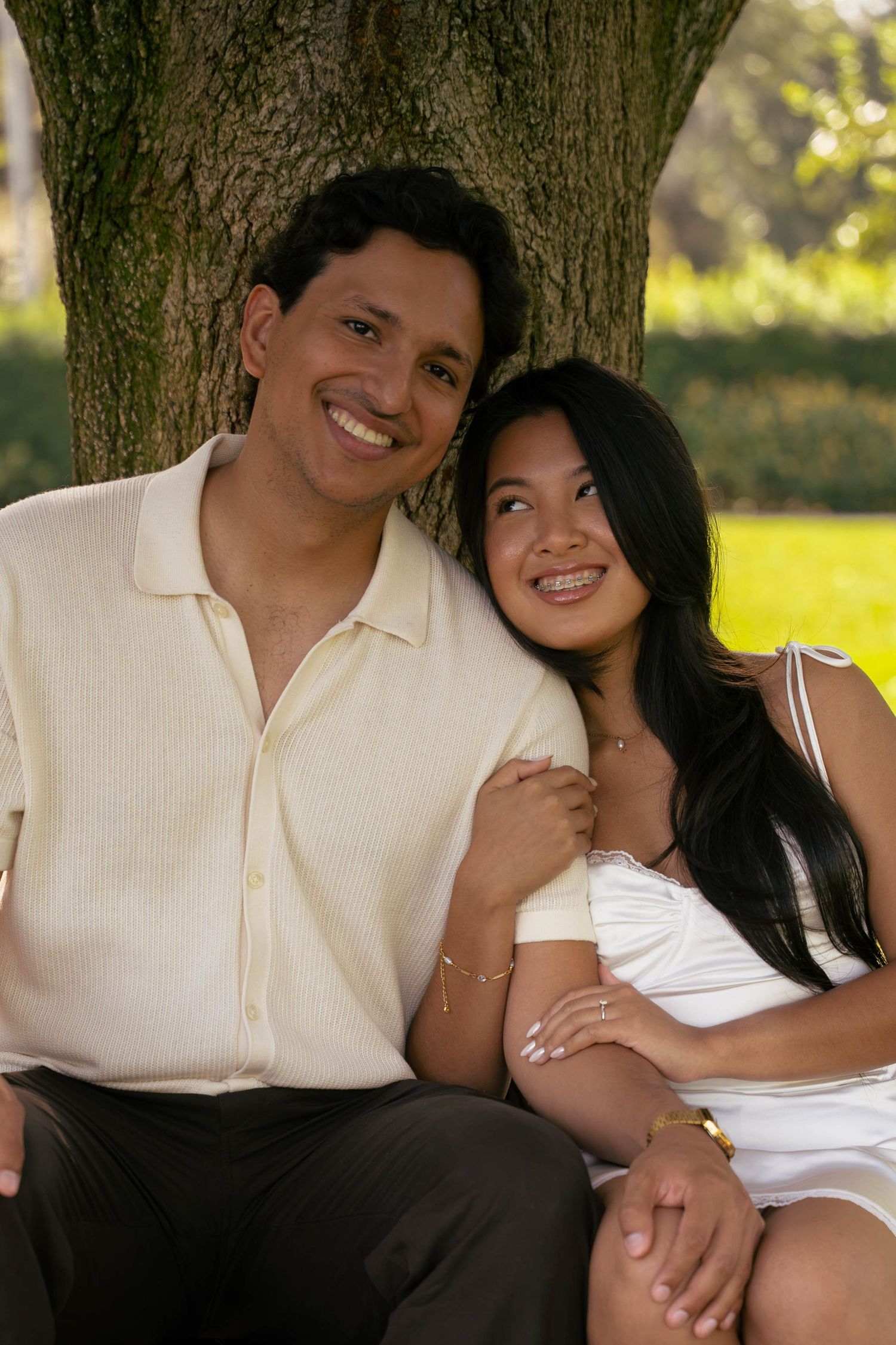 Smiling couple sitting together under a large tree in a sunny park setting.