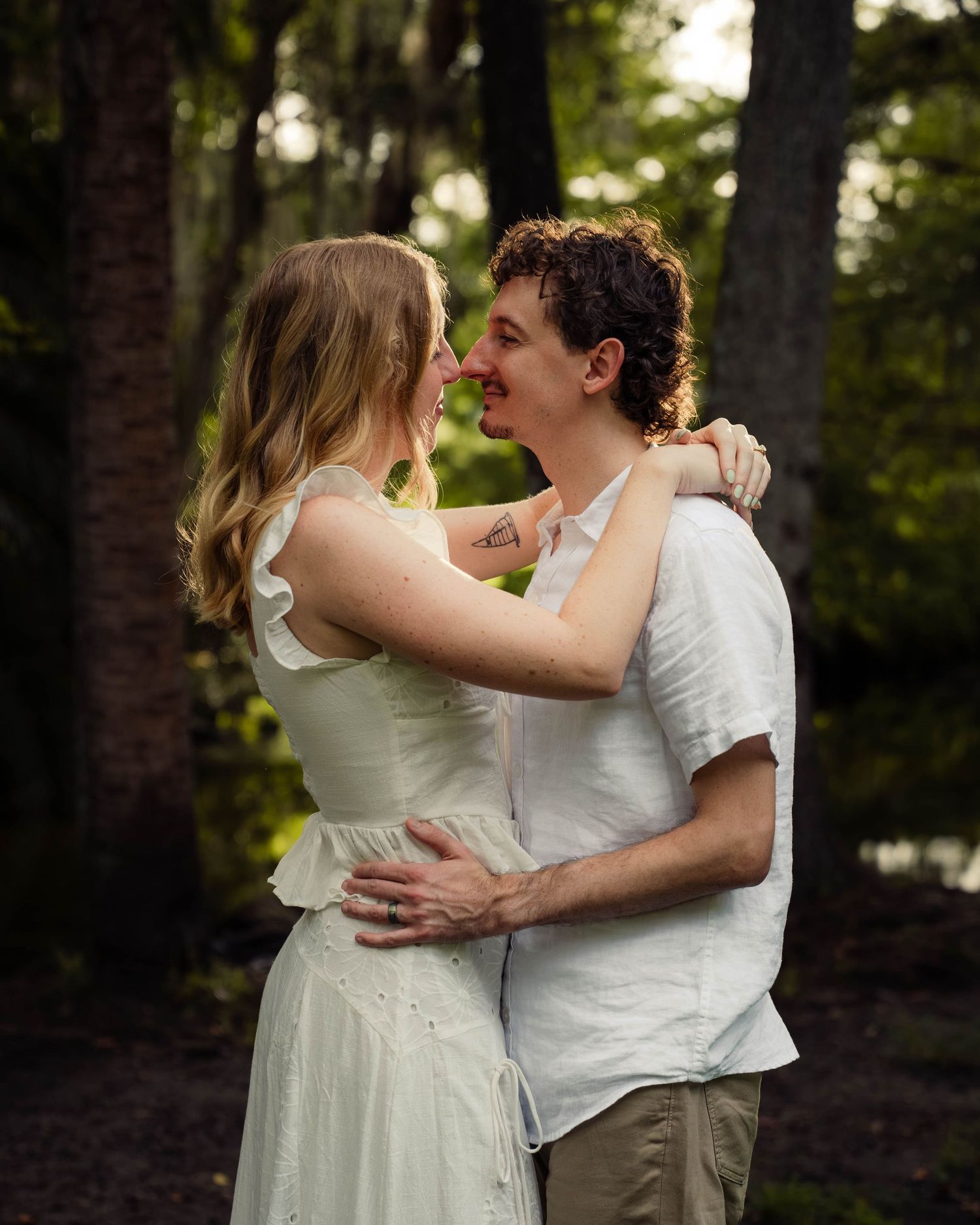 A couple embraces and smiles at each other among trees in a romantic outdoor setting.