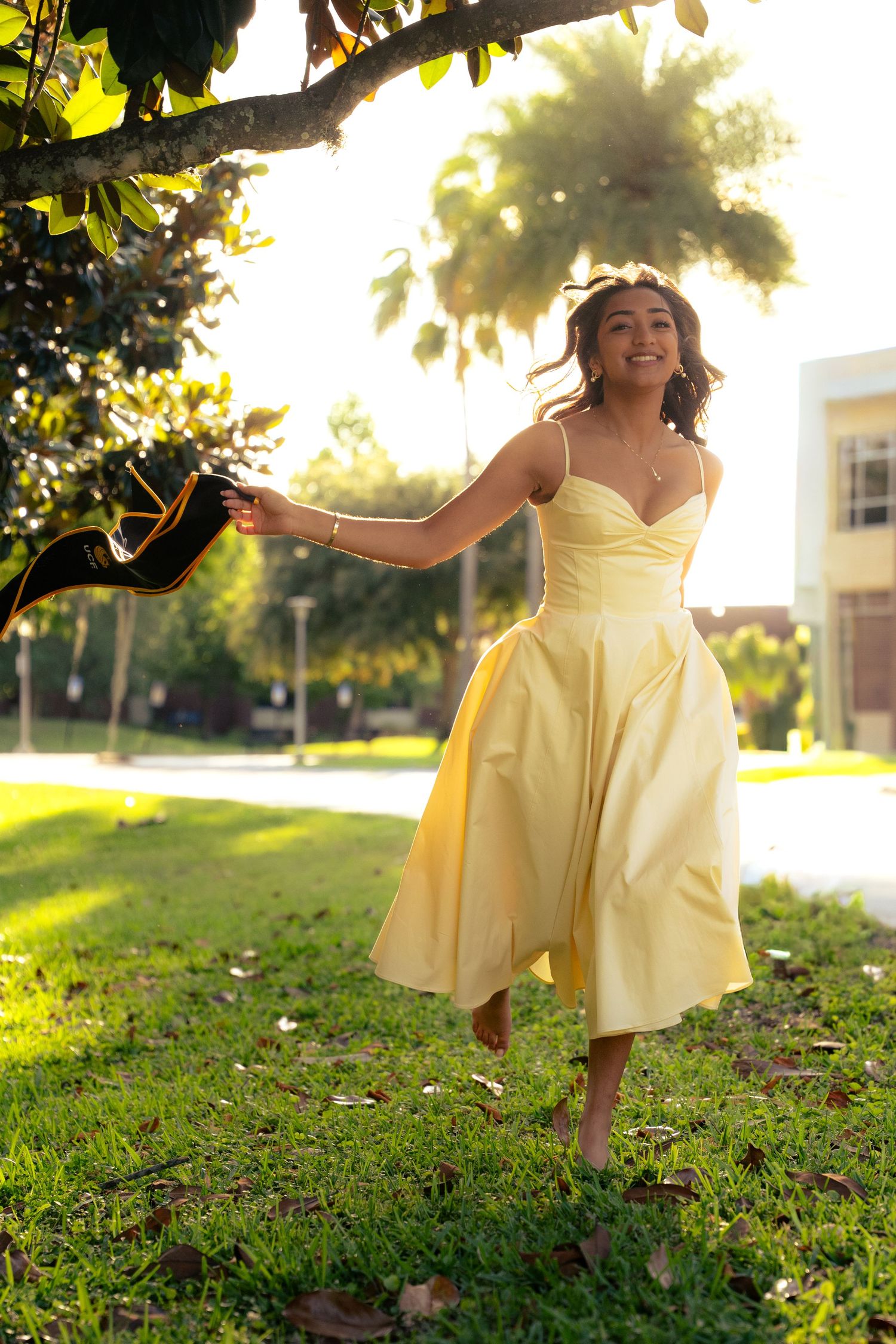 Smiling woman in yellow midi dress dances barefoot on grass holding heels in sunlight.